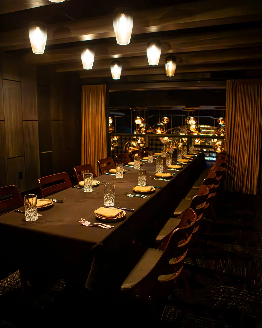 A dimly lit dining room with a long table set for a formal dinner, featuring glassware, plates, and napkins, with modern hanging lights and gold curtains in the background.