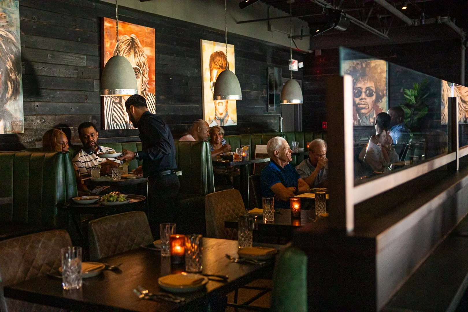 Dimly lit restaurant with diners seated in booths and at tables, decorated with colorful portrait artwork on black wood-paneled walls.