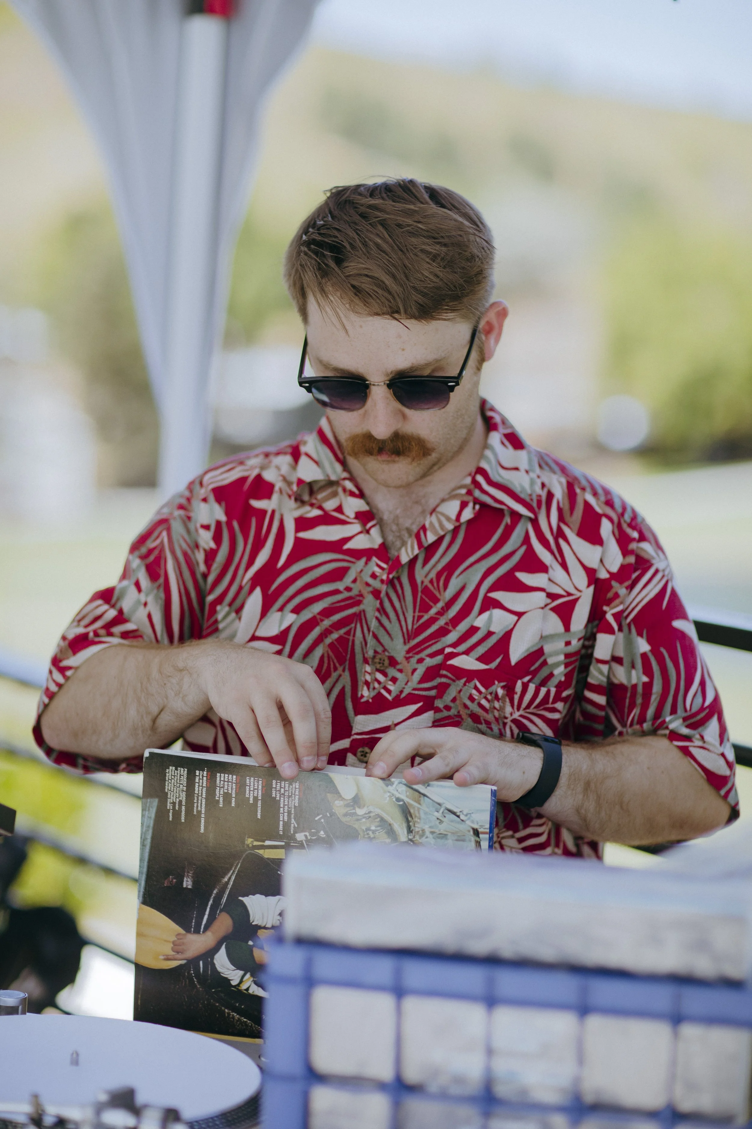 A man with brown hair, a mustache, wearing sunglasses and a red Hawaiian shirt, is spinning vinyl records for a DJ gig