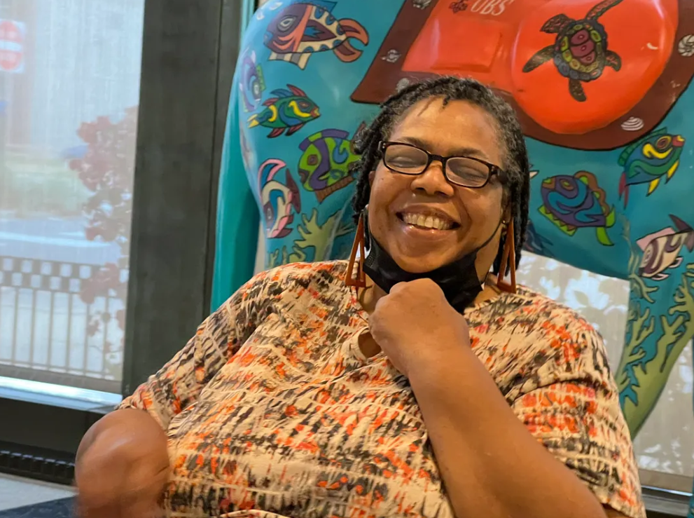 Smiling Black woman with glasses sits indoors in front of a colorful, fish-decorated sculpture. She wears a patterned shirt and a black mask under her chin.