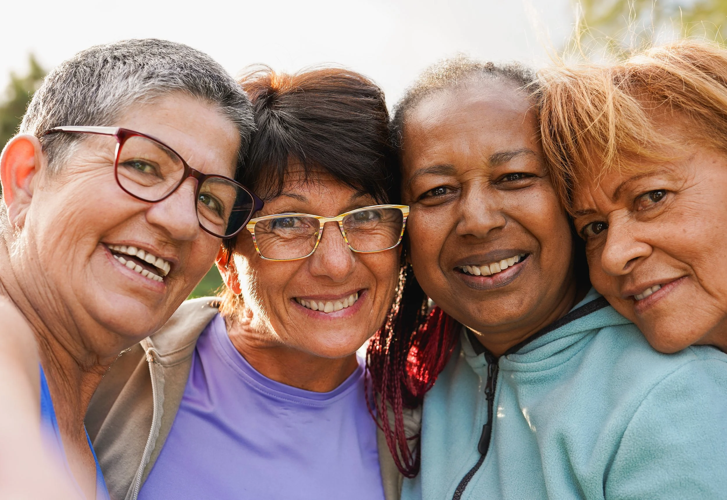 Four smiling older women of diverse backgrounds stand close together outdoors, taking a selfie. They are wearing casual clothes and glasses, and appear happy and relaxed in natural daylight.