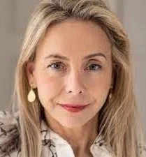 A white woman with long blonde hair, light skin, and wearing a white patterned blouse and teardrop-shaped earrings looks directly at the camera with a neutral expression.