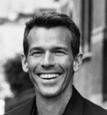 A white man with short hair smiles broadly while standing outdoors. The photo is in black and white, and the background is softly blurred, suggesting an urban street or building setting.