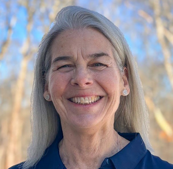 An older  white woman with long gray hair smiles outdoors, wearing a dark blue shirt and stud earrings. The background is blurred with leafless trees and clear blue sky.