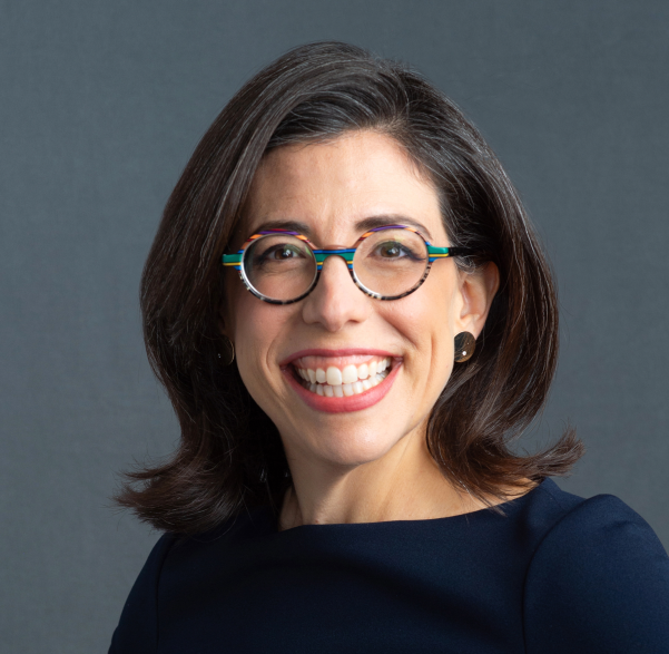 Smiling white woman with shoulder-length brown hair, wearing round multicolored glasses, black top, and stud earrings, in front of a plain gray background.