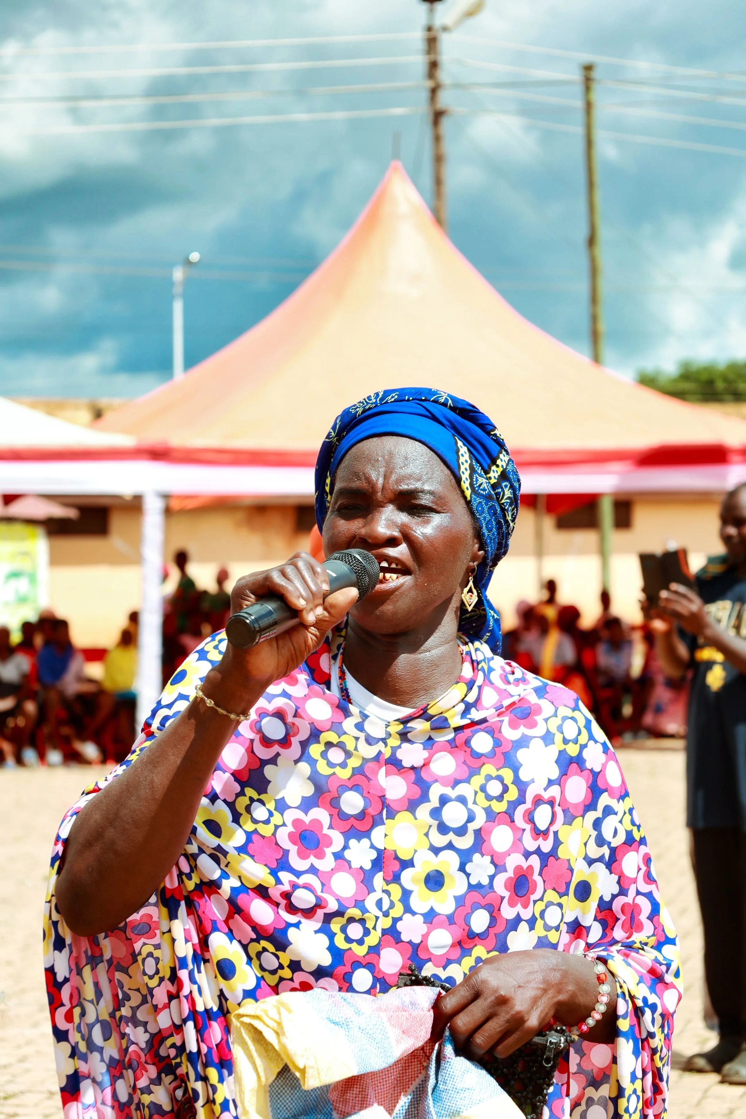 A dark-skinned woman in a colorful floral dress and blue headscarf speaks into a microphone outdoors, with people and tents in the background under a cloudy sky.