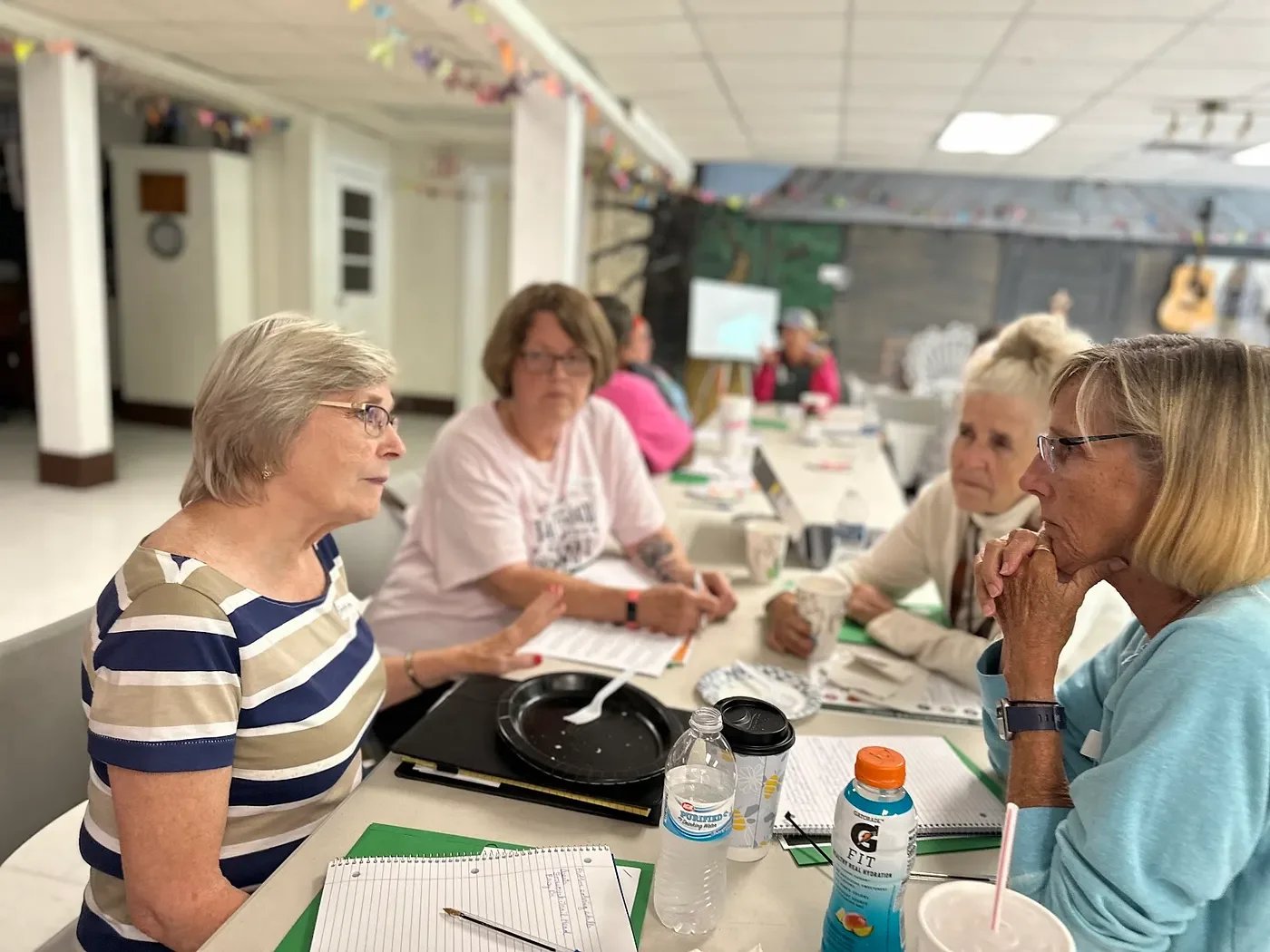 Four white women sit at a long table, engaged in conversation with notebooks, water, and snacks in front of them. The setting appears to be a casual group meeting in a community room.