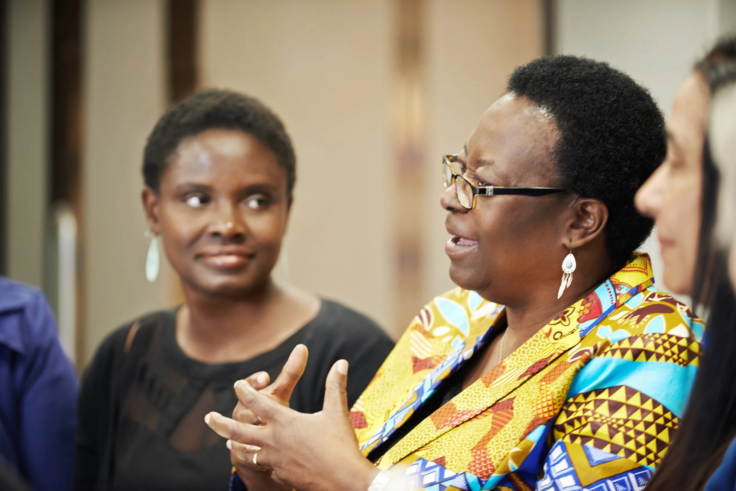 Two Black women are sitting together. One, wearing a colorful patterned outfit and glasses, is speaking while gesturing with her hands. The other woman listens attentively. The background is softly blurred.