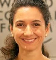 A light-skinned woman with dark hair tied back, wearing pearl earrings, smiles at the camera. The background is blurred with partial, indistinct text visible.