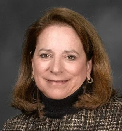 A professional headshot of a light-skinned woman with shoulder-length brown hair, smiling gently at the camera. She is wearing a black turtleneck under a textured, dark-patterned blazer against a simple gray background.