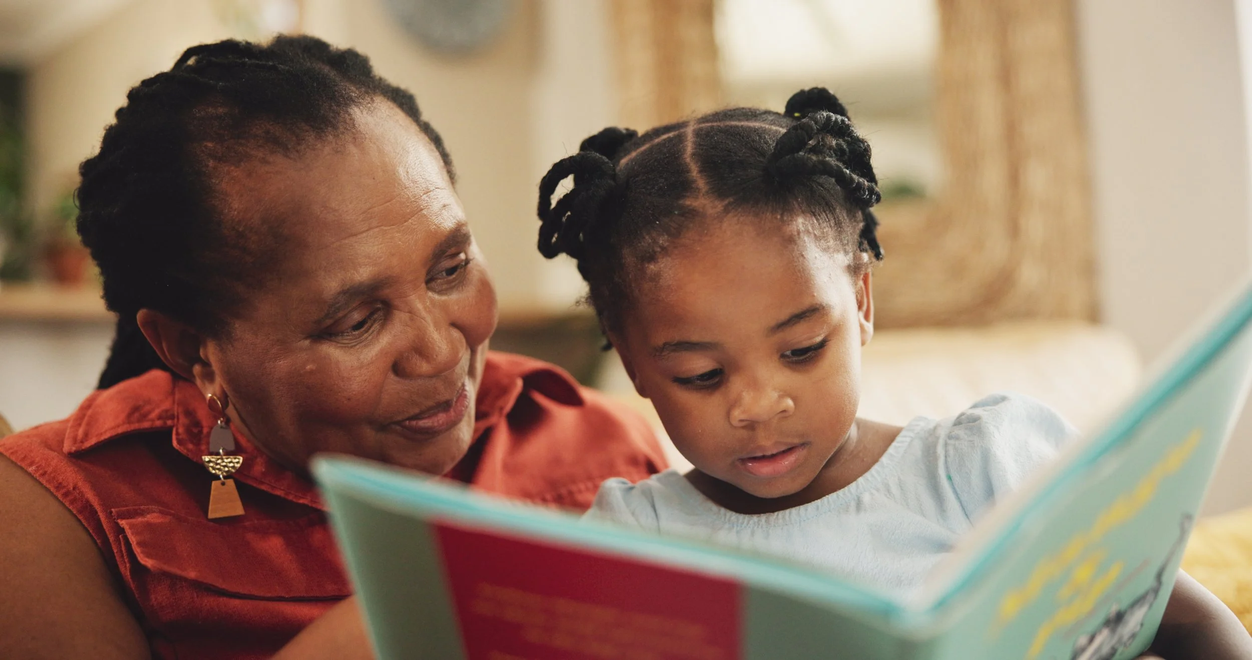 An older Black woman and a young Black girl sit closely together on a couch, reading a colorful childrens book. The woman smiles gently while the girl looks intently at the pages.