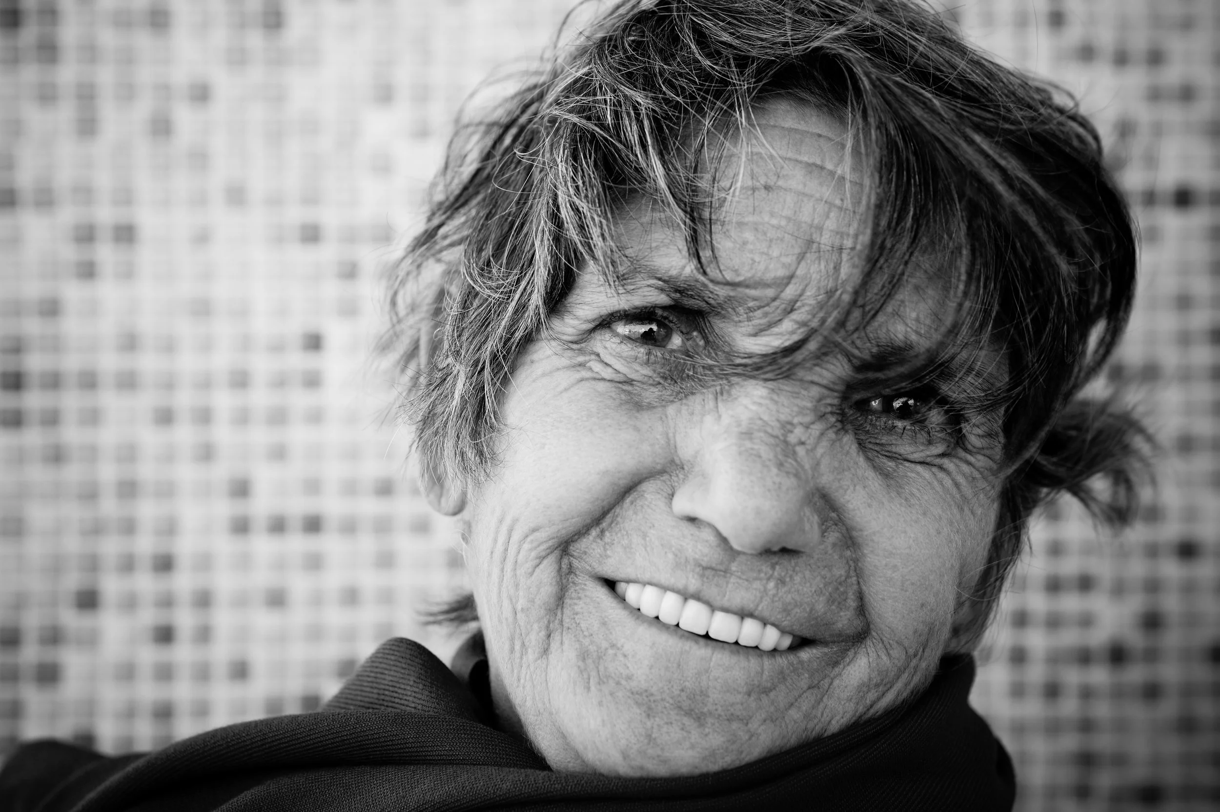 Smiling older woman with tousled hair, wearing a dark top, poses in front of a blurred mosaic tile background. The photo is in black and white, emphasizing her facial features and joyful expression.