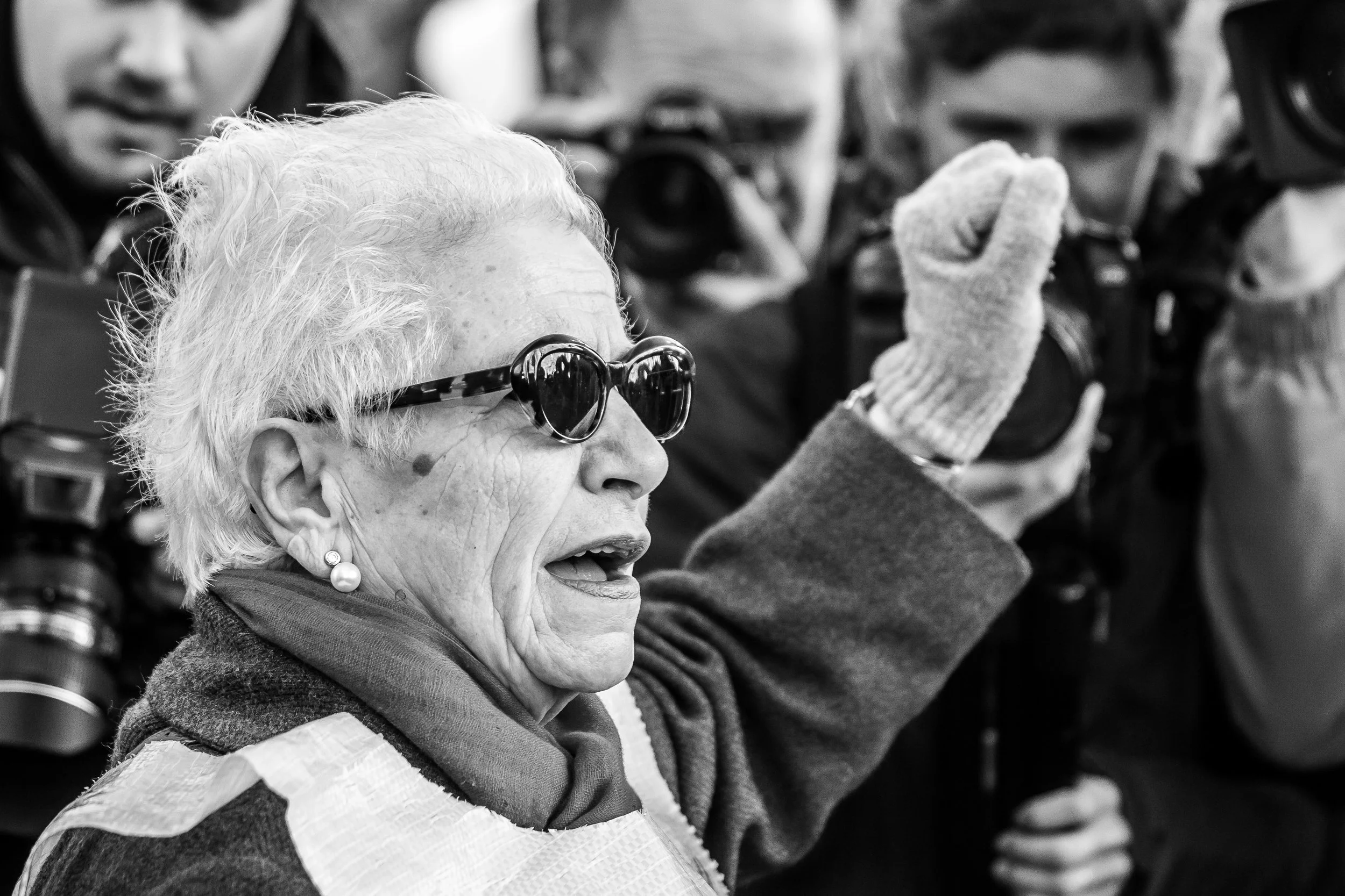 An older woman wearing sunglasses raises her gloved fist while speaking. Photographers with cameras surround her, capturing the moment. The image is in black and white.