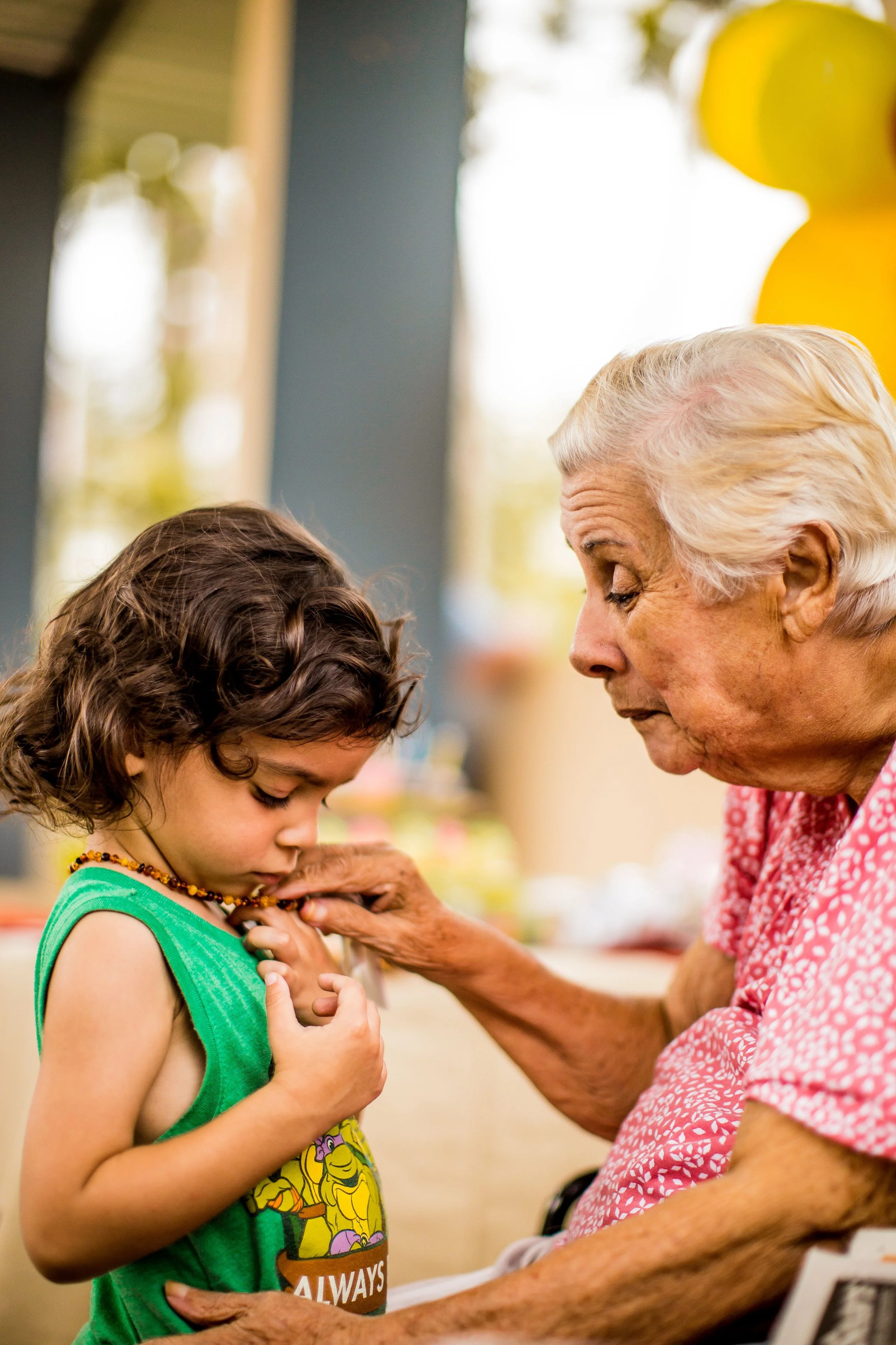 An elderly woman gently helps a young child adjust a necklace. The child looks down at the necklace, and both share a tender moment.
