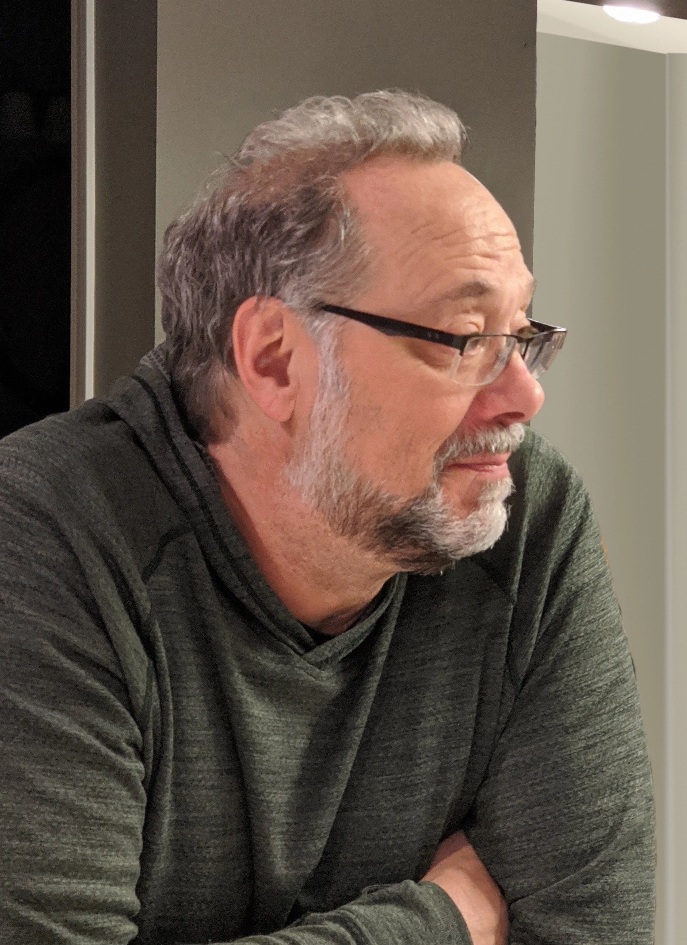 Side profile of the artist wearing glasses, and a dark green hoodie with a black zipper, sitting at a table with his arms crossed.