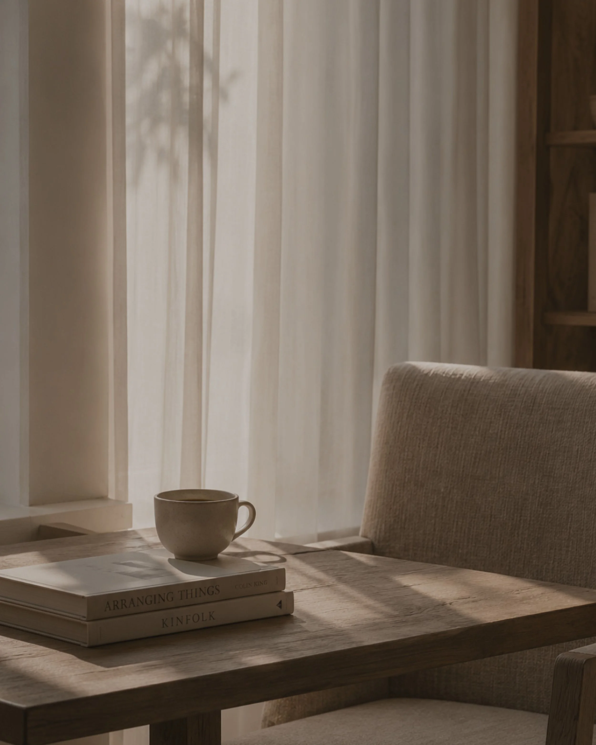 A cozy interior scene showing a wooden table with three books, a white ceramic mug, a window with sheer curtains, a beige upholstered chair, and wooden shelves in the background.