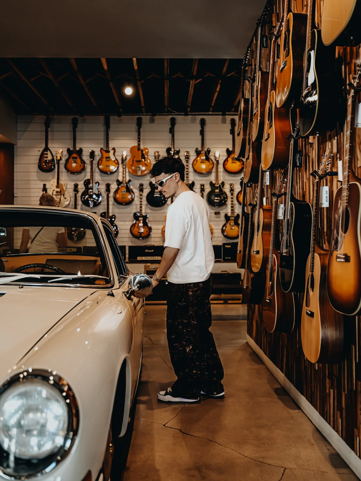 A man with dark hair, wearing sunglasses, a white t-shirt, and black pants, stands beside a vintage cream-colored car inside a guitar store. The store has a wall of hanging guitars behind him and guitars on a wooden wall to the right.