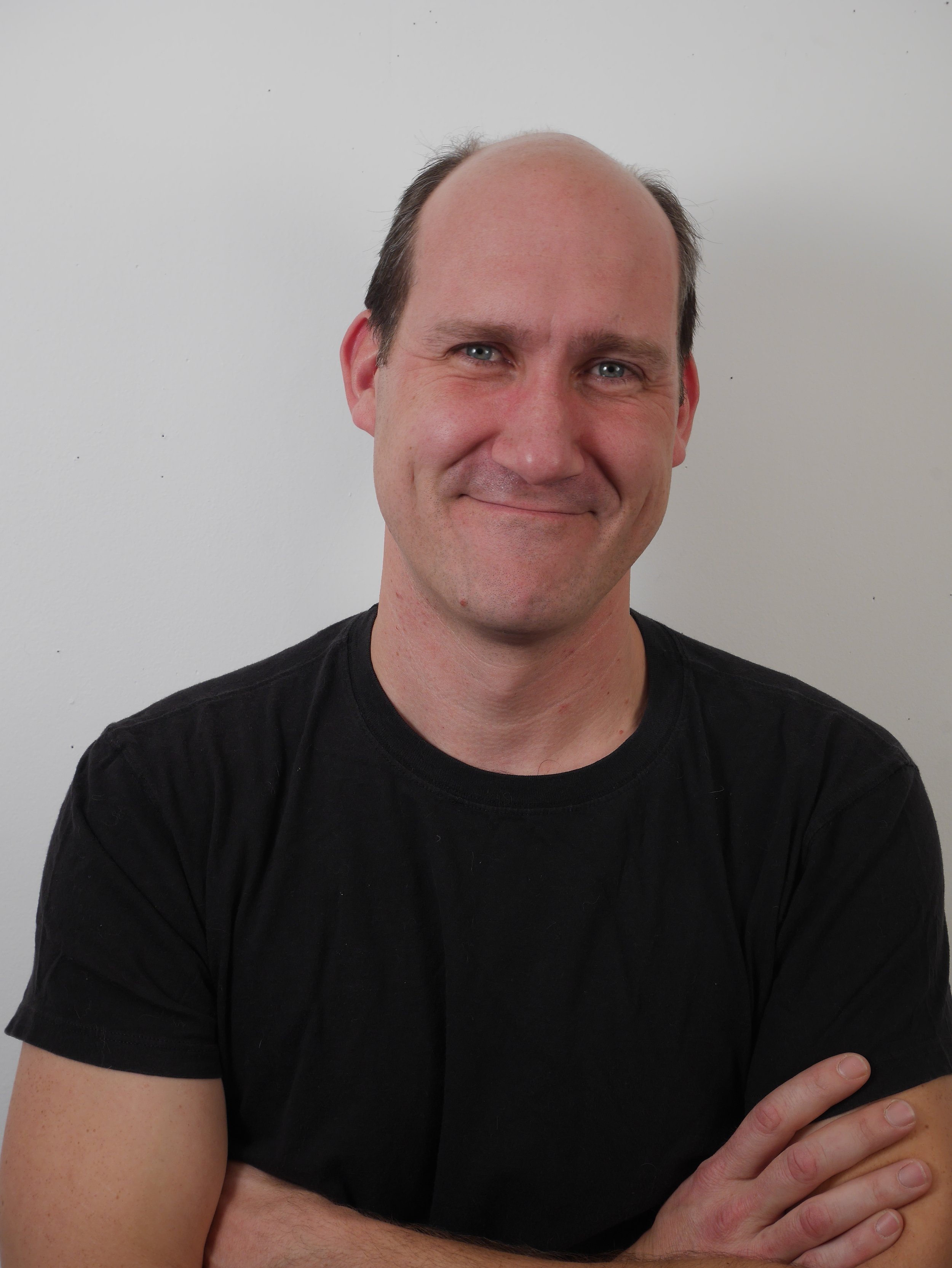 Close-up photo of a smiling man with a receding hairline, wearing a black shirt, standing against a plain white background.