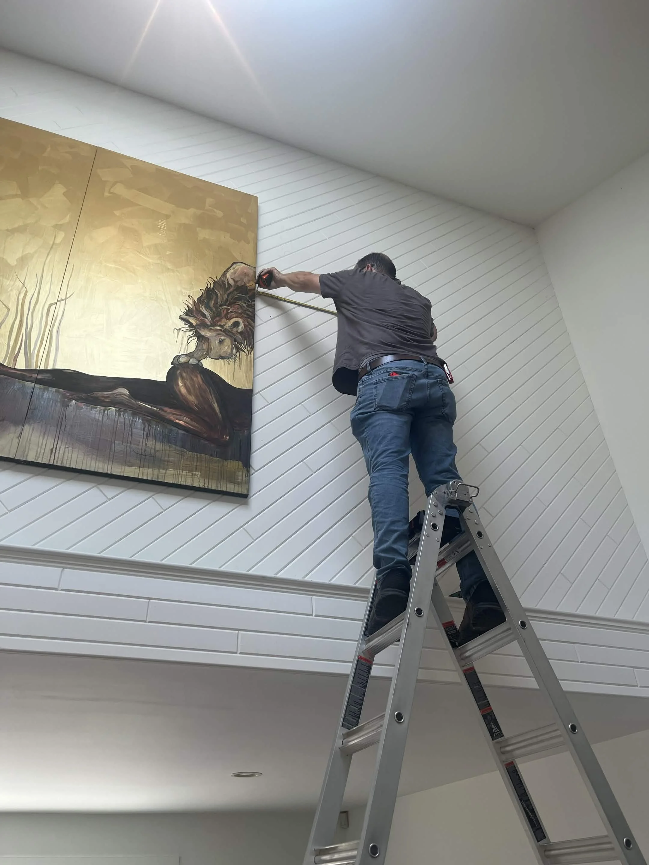 A man standing on a ladder installing or working on a large framed painting of a lion in an indoor space, with white and light gray walls.
