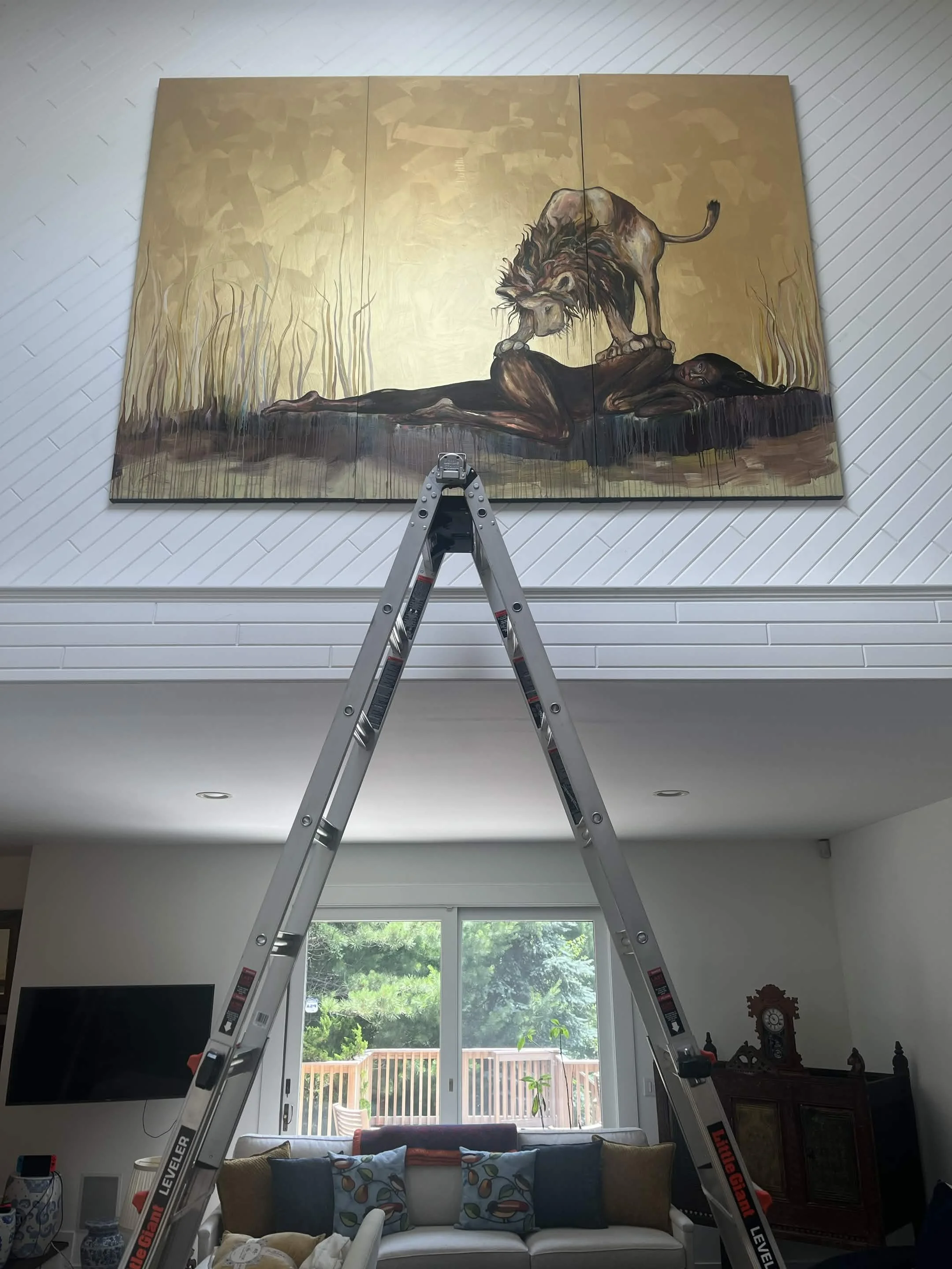 Interior of a living room with a large series of three painting panels depicting a lion standing over a reclining woman. The painting is on a wall above a beige sofa with colorful pillows. In front, a silver ladder is positioned, and large windows at the back provide natural light and a view of greenery outside.