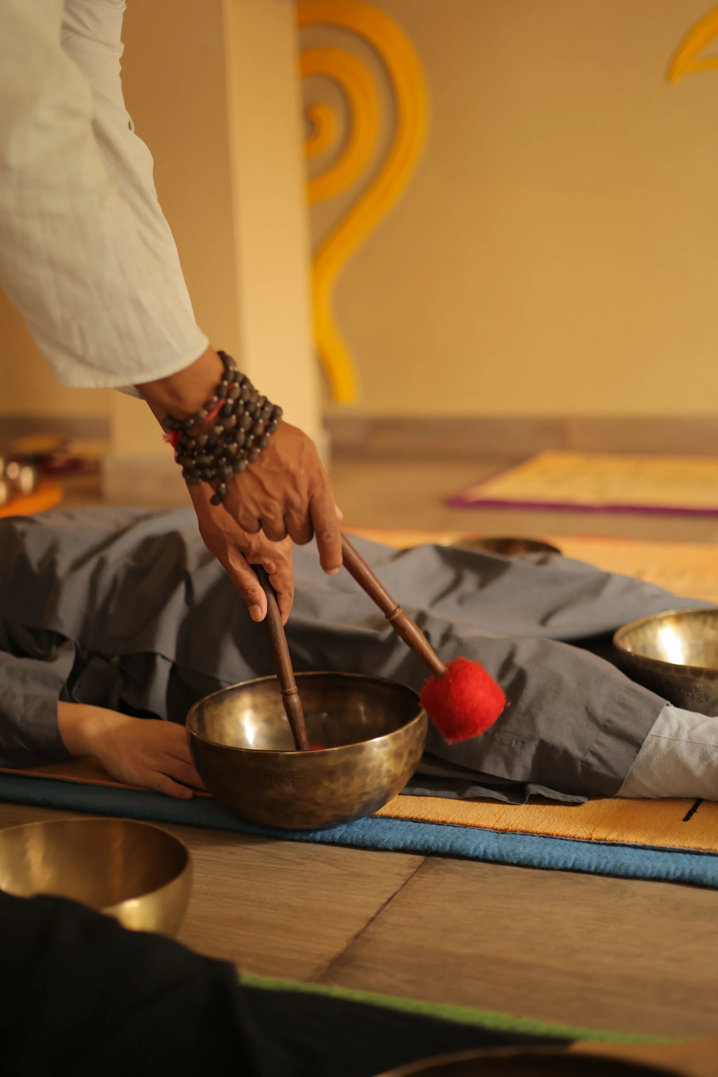 Orin App person wearing a white garment and beaded bracelets uses a wooden mallet to strike a singing bowl during a spiritual or meditation session.