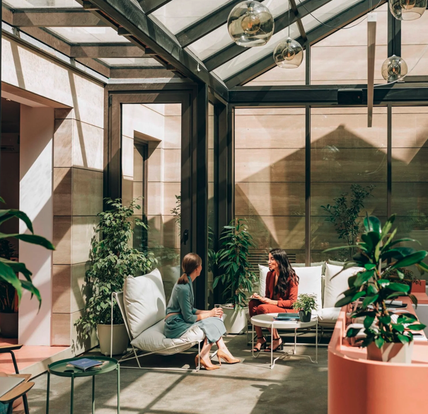 Two women chatting in a modern indoor lounge with large glass windows, white seating, and plants.