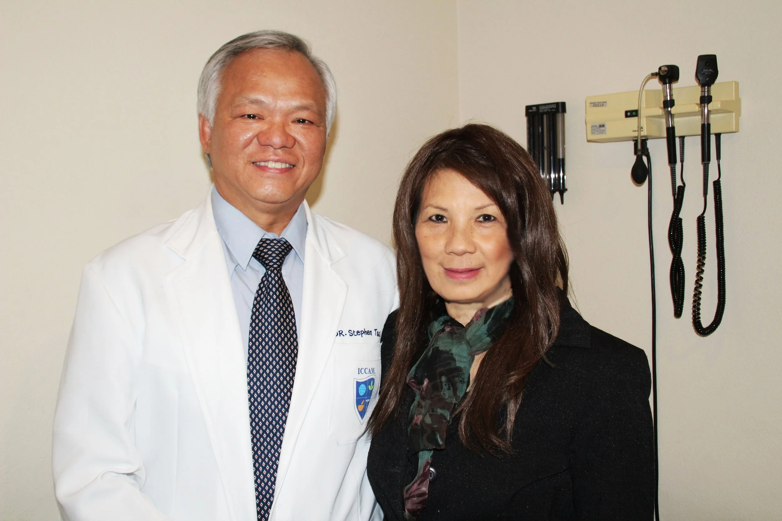 A doctor and a woman posing together in a medical office, with medical equipment on the wall behind them.