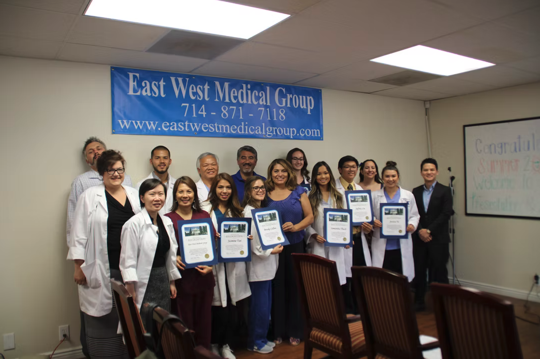 Group of people, including medical professionals and students, holding certificates during a ceremony at East West Medical Group office.