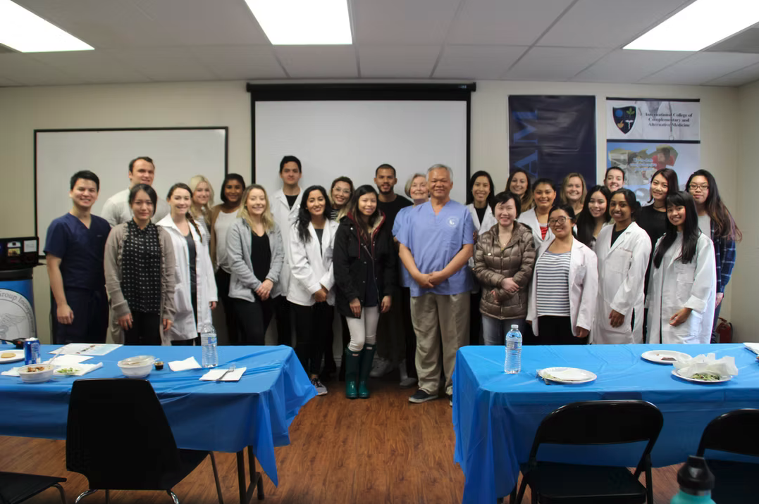 Group of students and a professor posing in a classroom or conference room with tables set with plates, water bottles, and food.