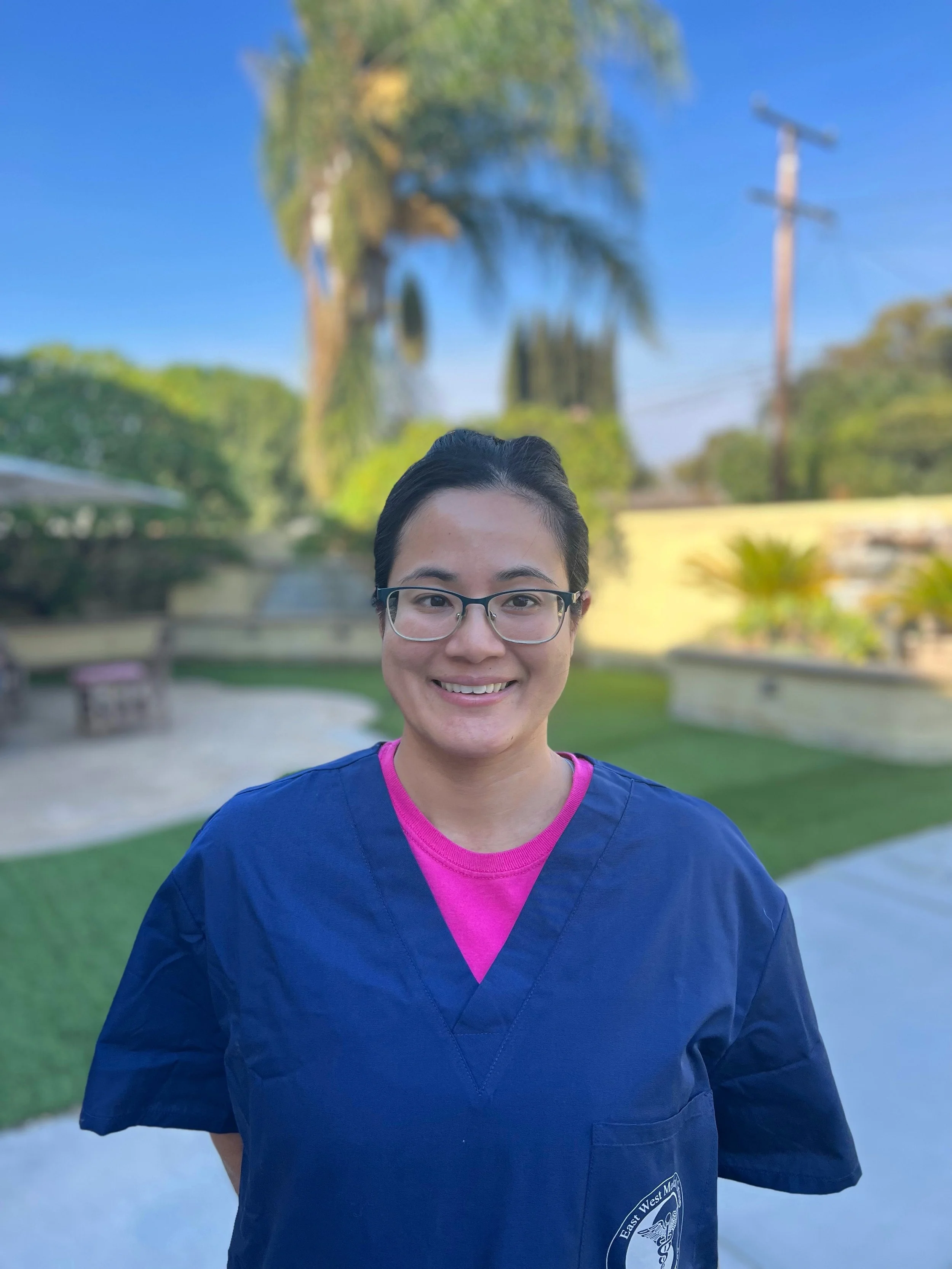 A person wearing glasses and a navy blue medical scrubs top with pink inner shirt, smiling outdoors with palm trees, a utility pole, and a bright blue sky in the background.