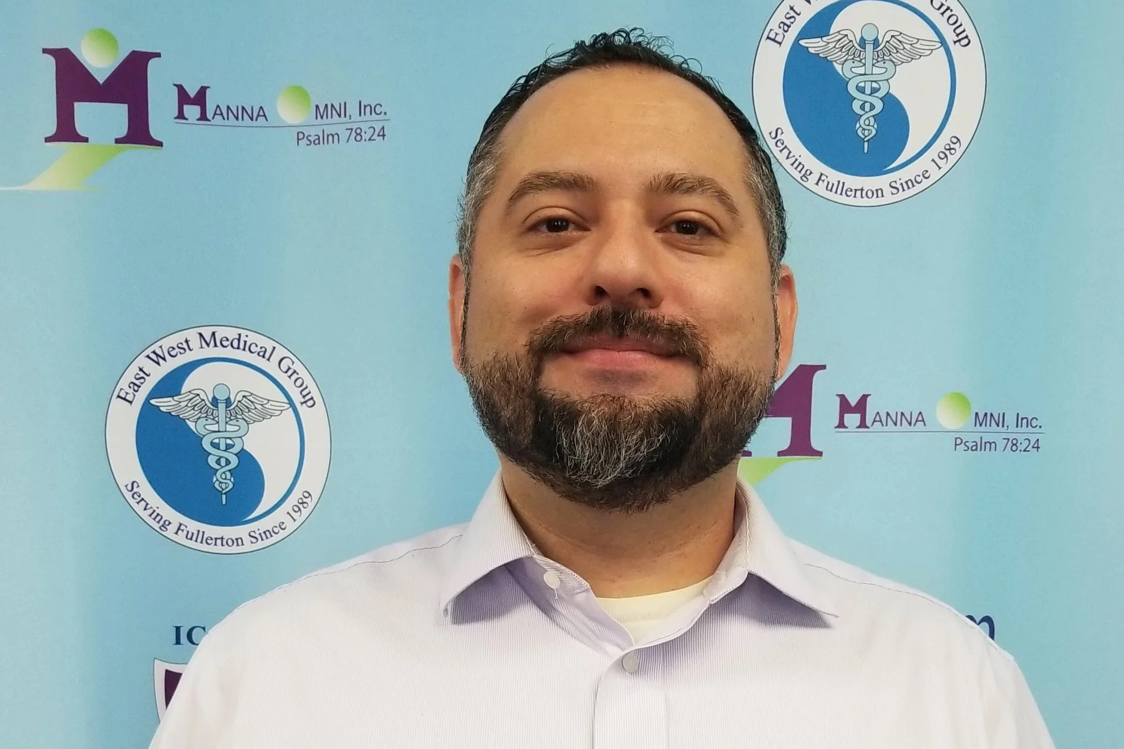 A man with dark hair and a beard smiling, wearing a light purple collared shirt, standing in front of a light blue background with medical and organization logos.