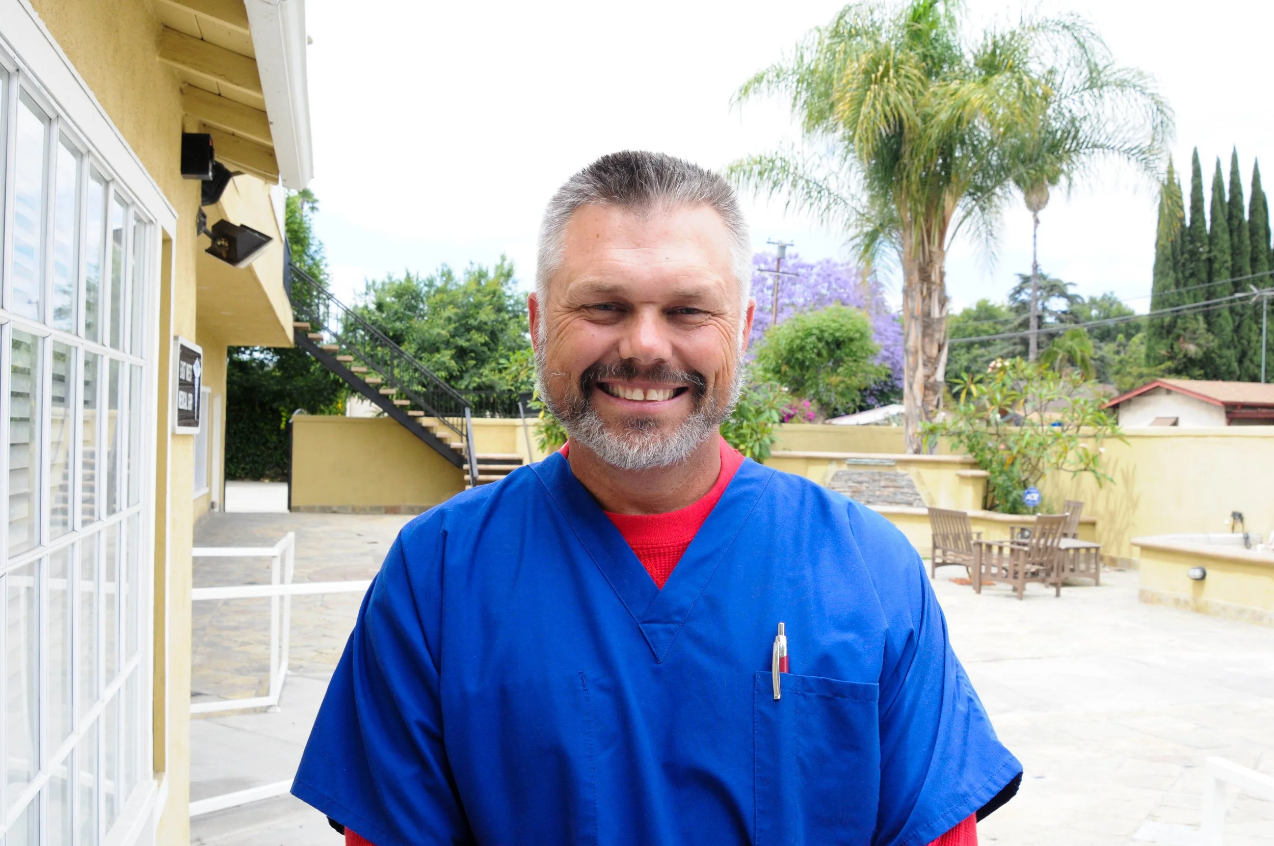 Smiling man in blue scrubs outdoors in a backyard with palm trees and purple flowering trees.