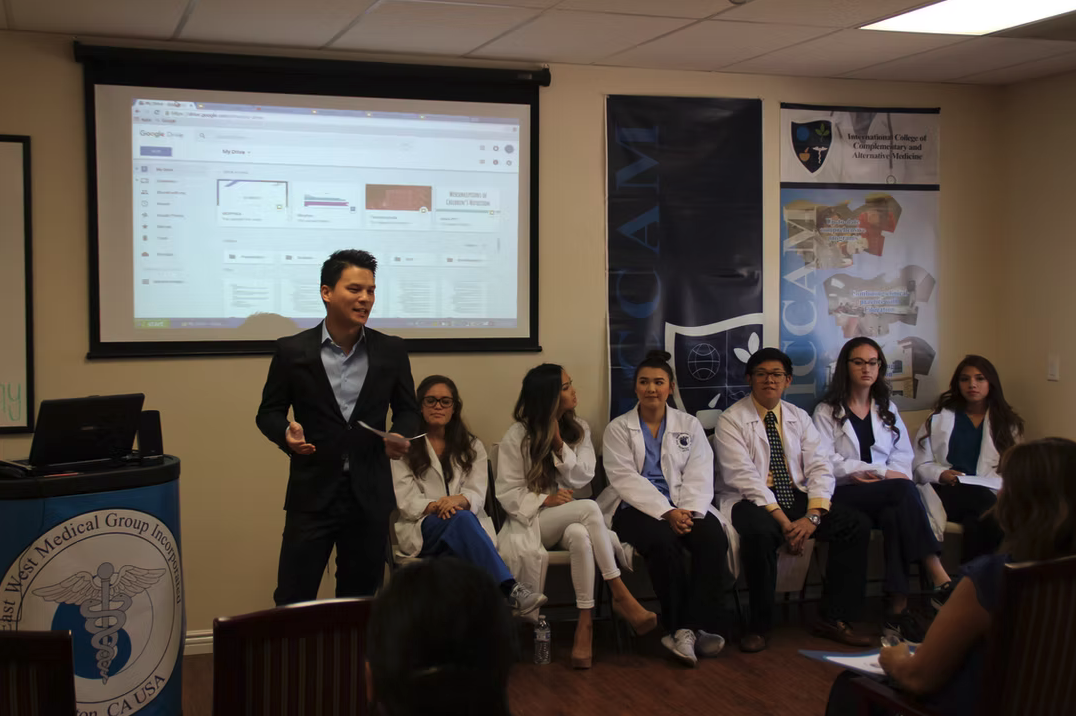 A group of medical students, some in white coats, sitting on a bench in a classroom. A man in a suit is standing and speaking, holding notes. Behind them, a large screen displays a computer interface and banners with medical school logos.