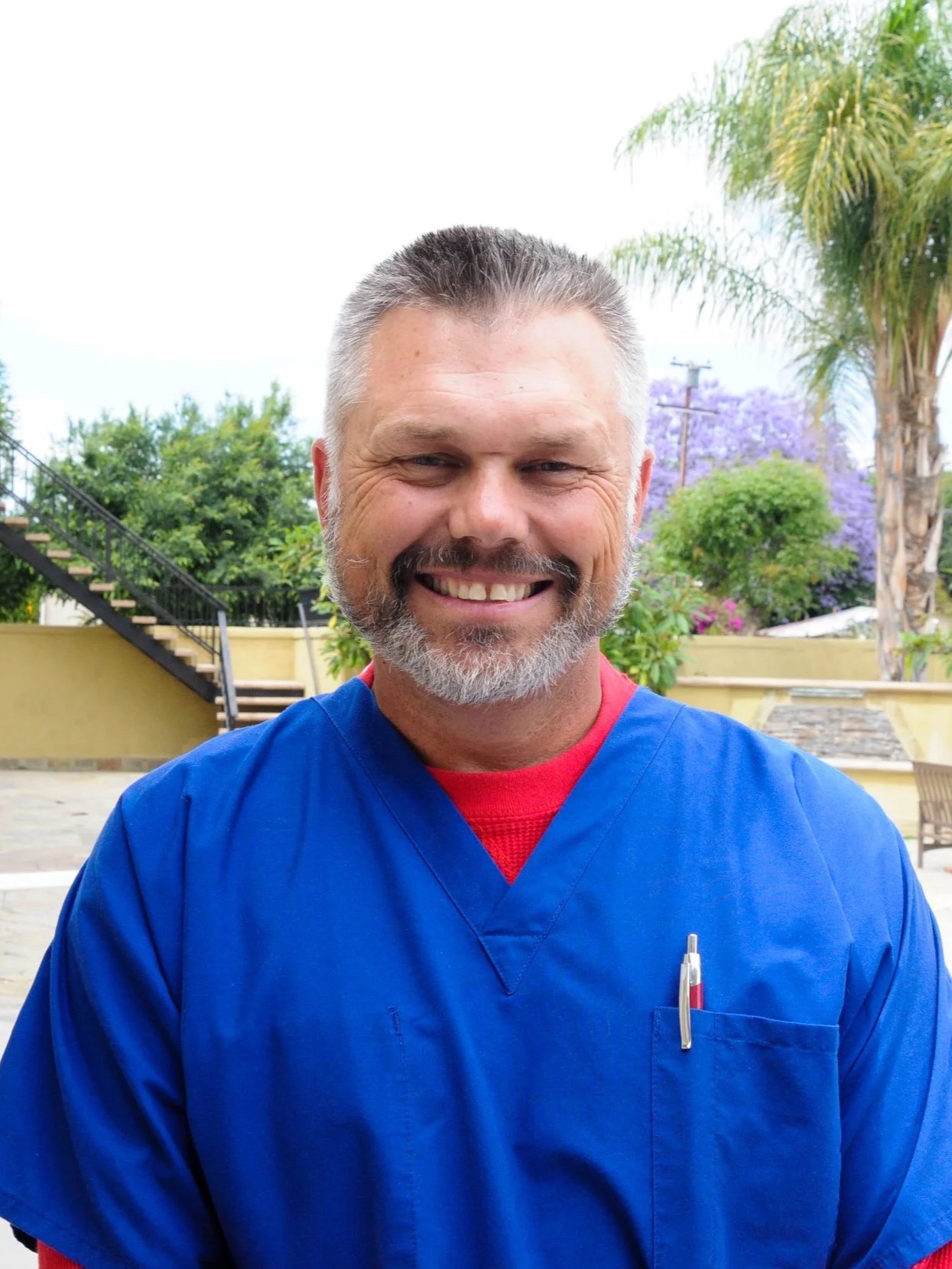 A smiling male healthcare professional with gray hair and a beard, wearing blue scrubs and a red undershirt, outdoors with trees and plants in the background.