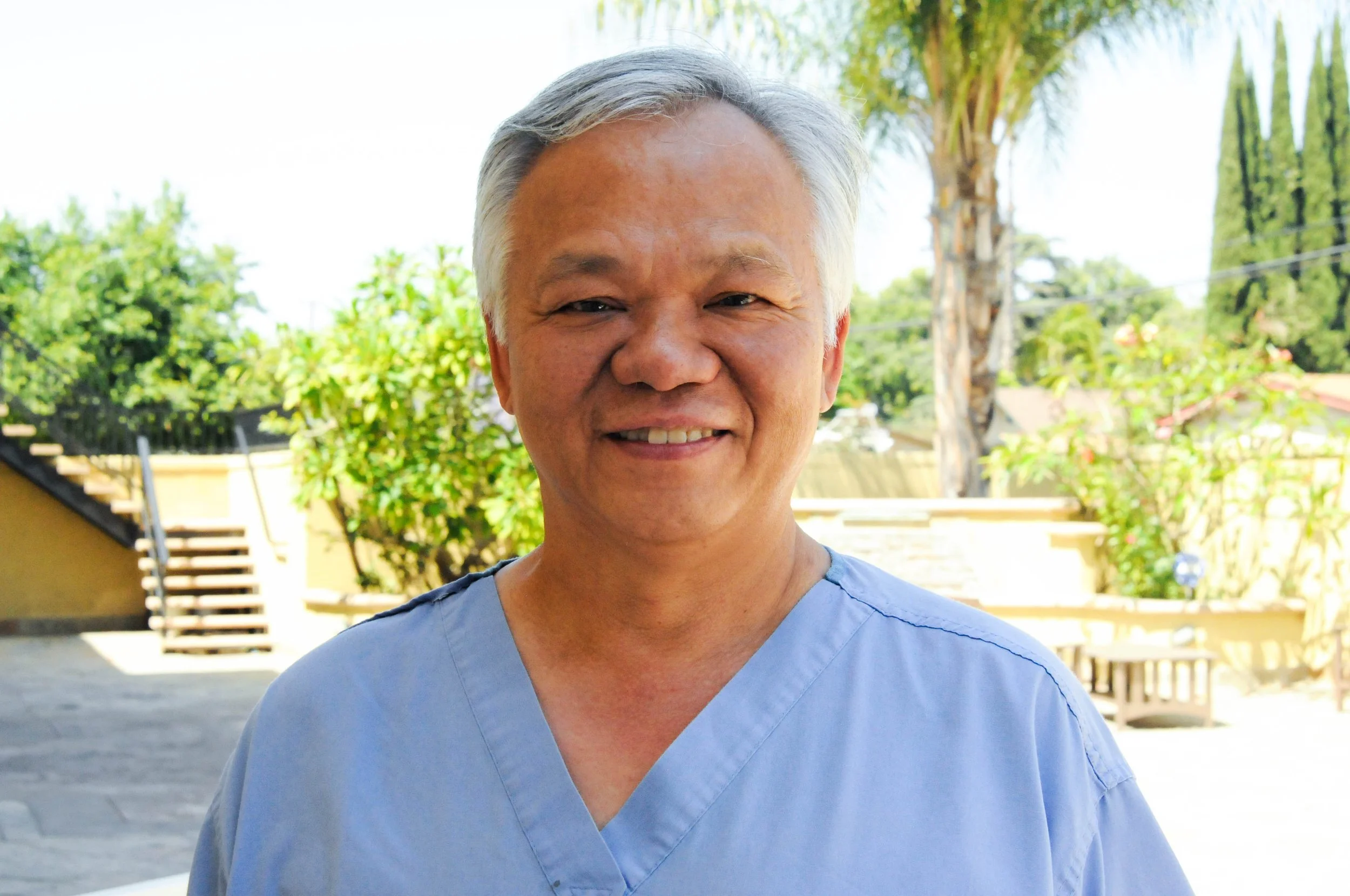 A middle-aged man with gray hair wearing light blue medical scrubs, smiling outdoors with green trees and a wooden fence in the background.