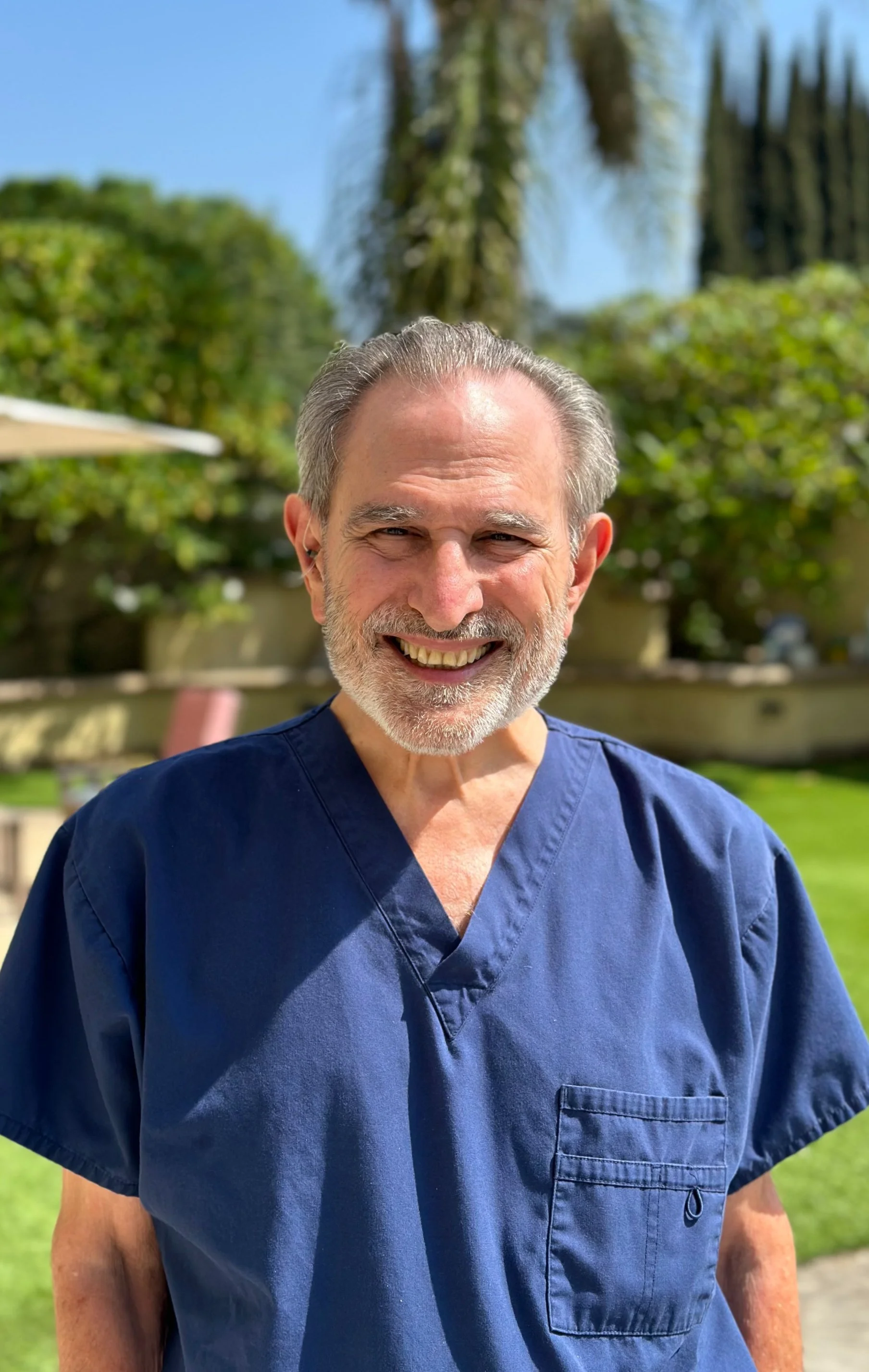 A man with gray hair and beard smiling outdoors in a nurse or medical scrubs uniform.