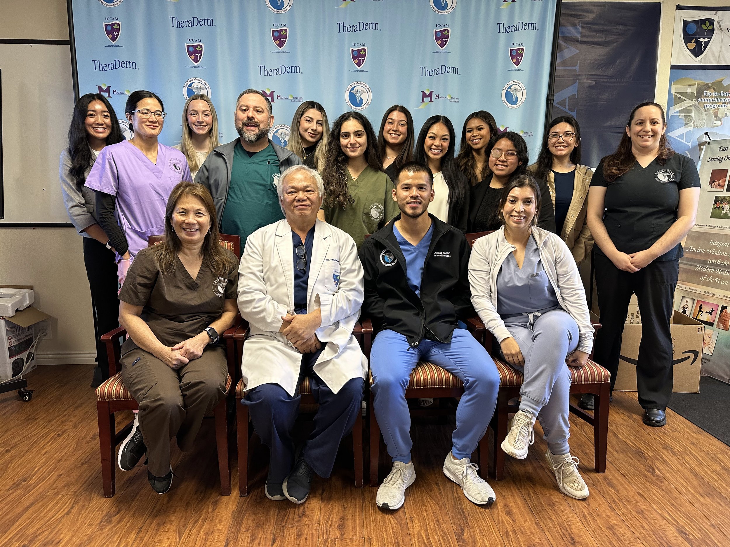 A group of diverse medical professionals and students posing for a photo in front of a blue backdrop with logos and text. They are smiling and sitting or standing together.