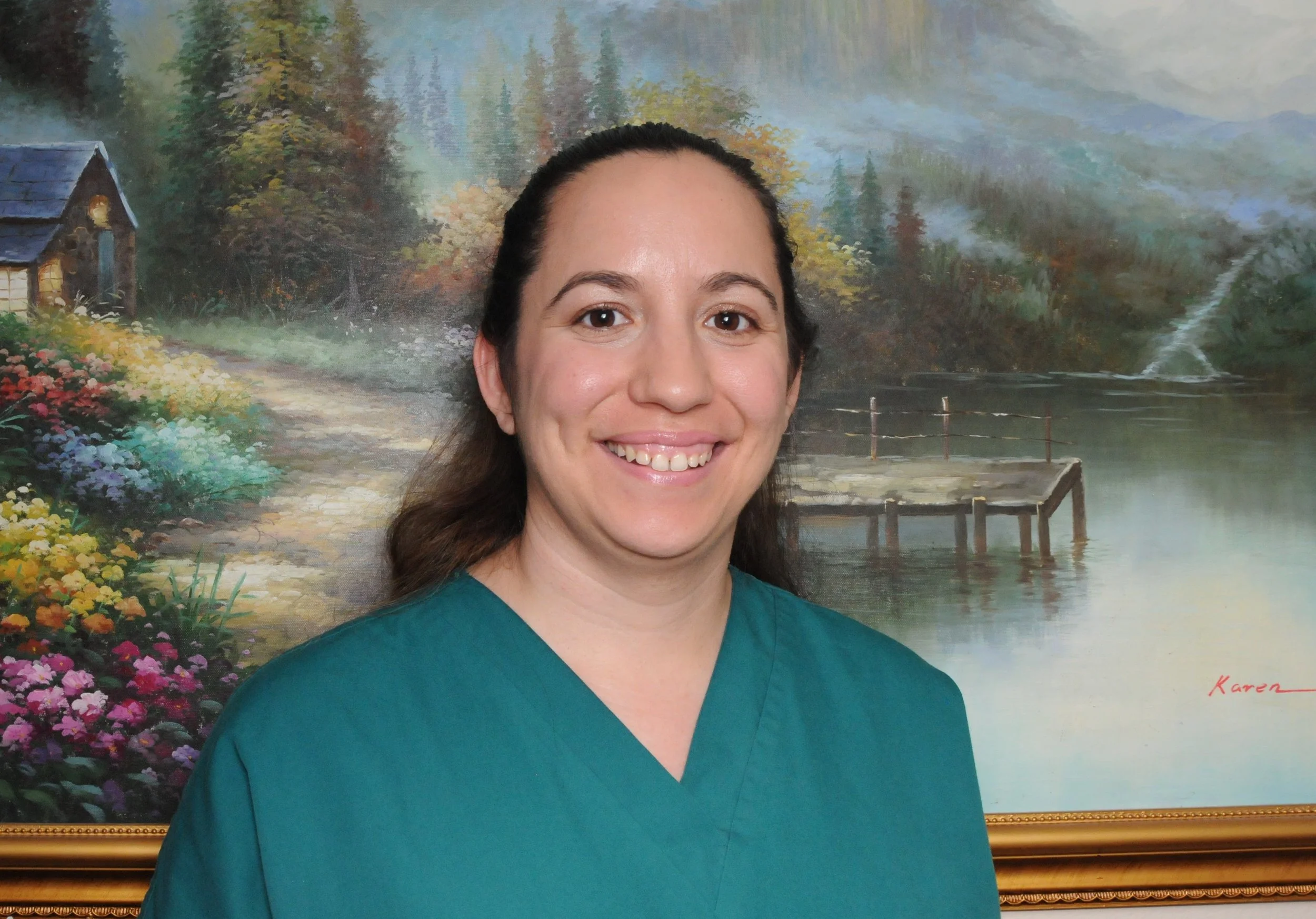 A woman with long brown hair, smiling, wearing a teal medical uniform, standing in front of a large landscape painting of a serene lake, trees, and mountain scenery.
