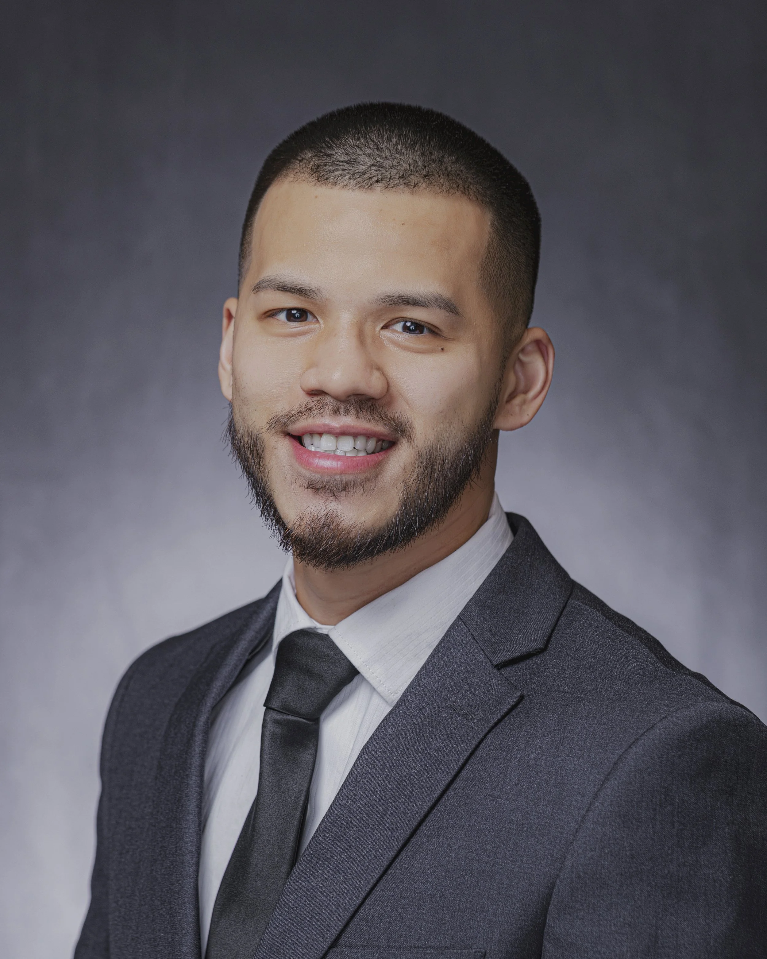 Close-up portrait of a smiling man wearing a suit and tie, with short dark hair and a beard, against a gray gradient background.