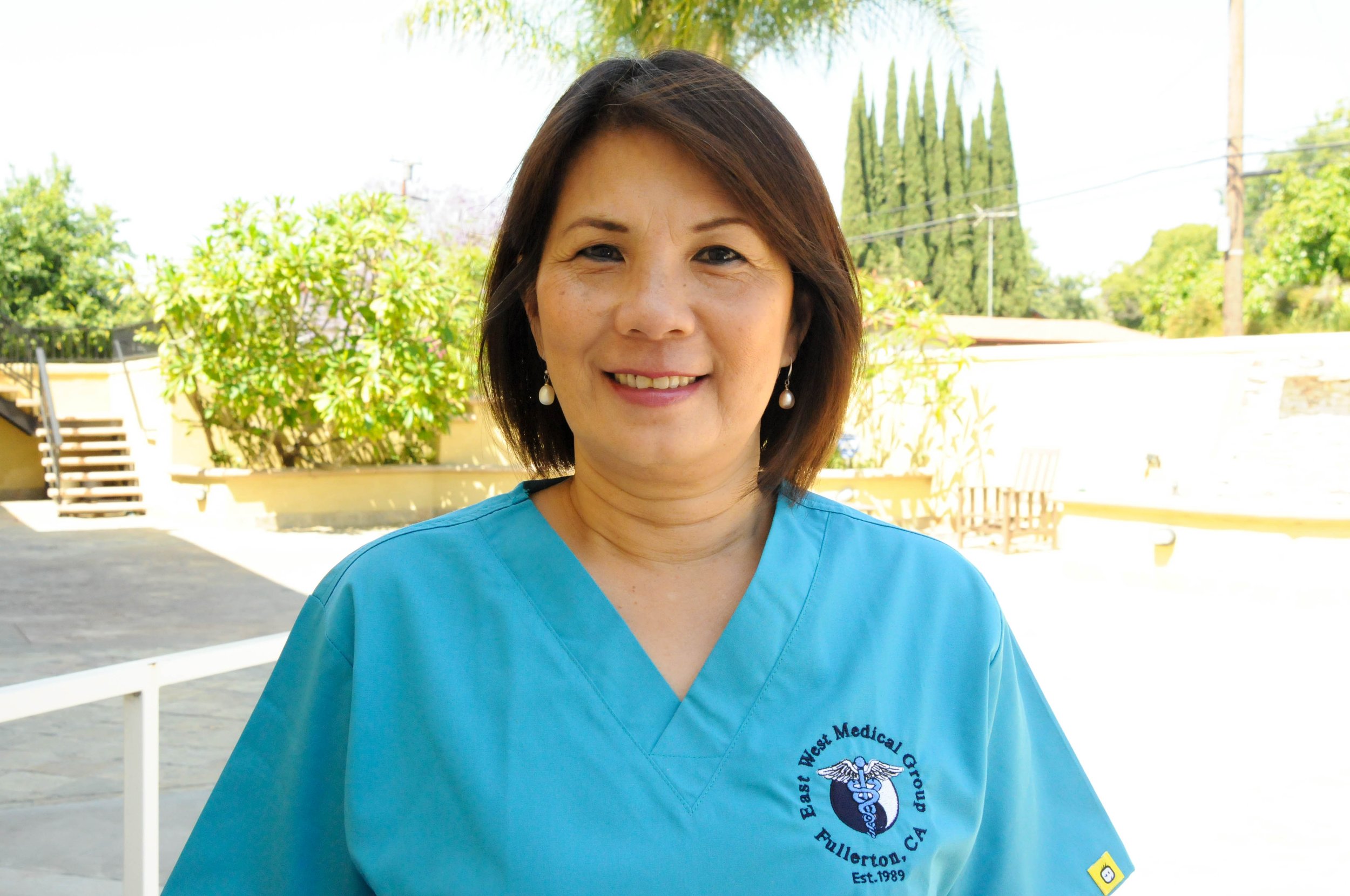 A woman in medical scrubs standing outdoors with greenery and trees in the background.