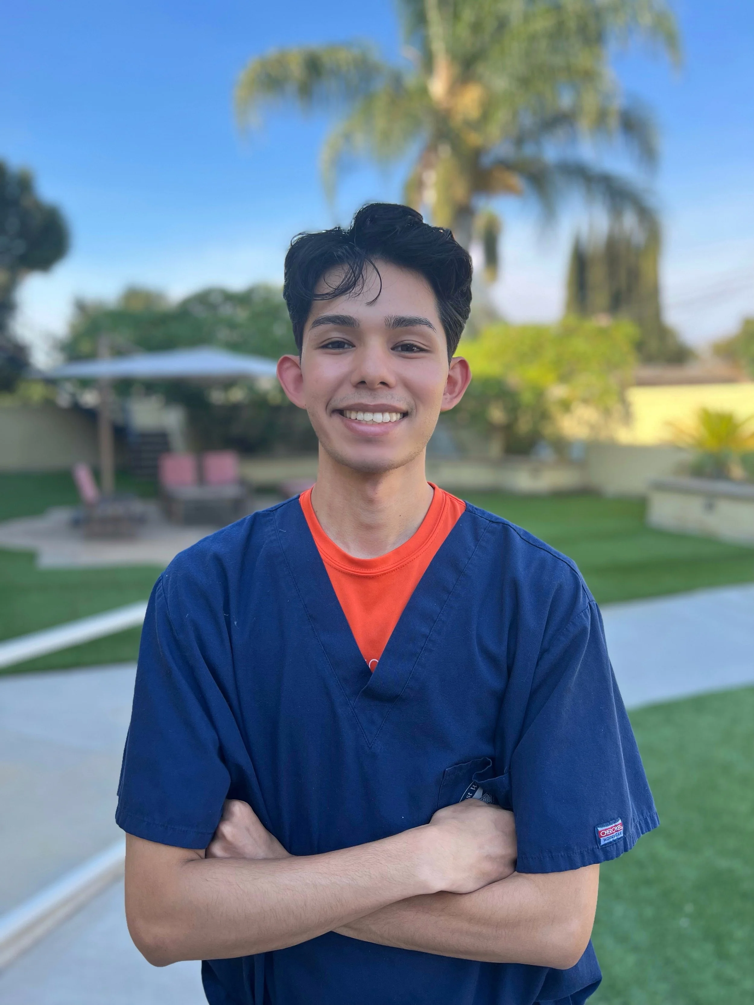 A young man with dark hair and a smile, wearing a navy blue scrub top over an orange shirt, standing outdoors with arms crossed, background featuring a palm tree, a lawn, and a clear blue sky.