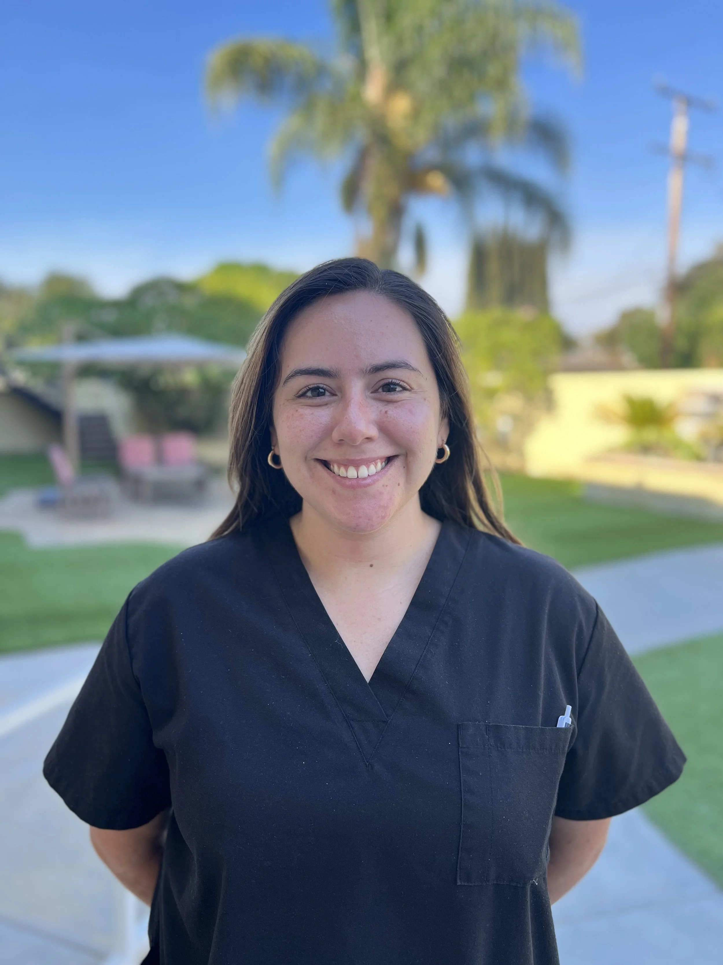 A smiling woman in a black medical uniform standing outdoors in front of a park with a palm tree and clear blue sky.