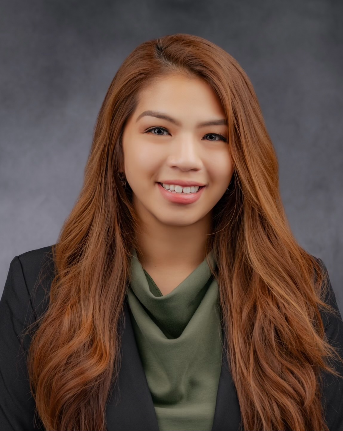 Portrait of a woman with long, wavy, reddish-brown hair, smiling, wearing a black blazer and an olive green shirt, against a dark gray background.