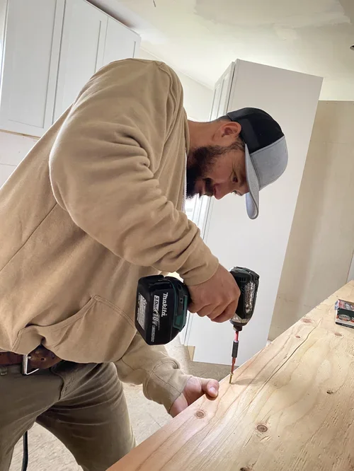 A man wearing a beige hoodie and a baseball cap, using a cordless drill, is working on a wooden surface in a room with white cabinets.