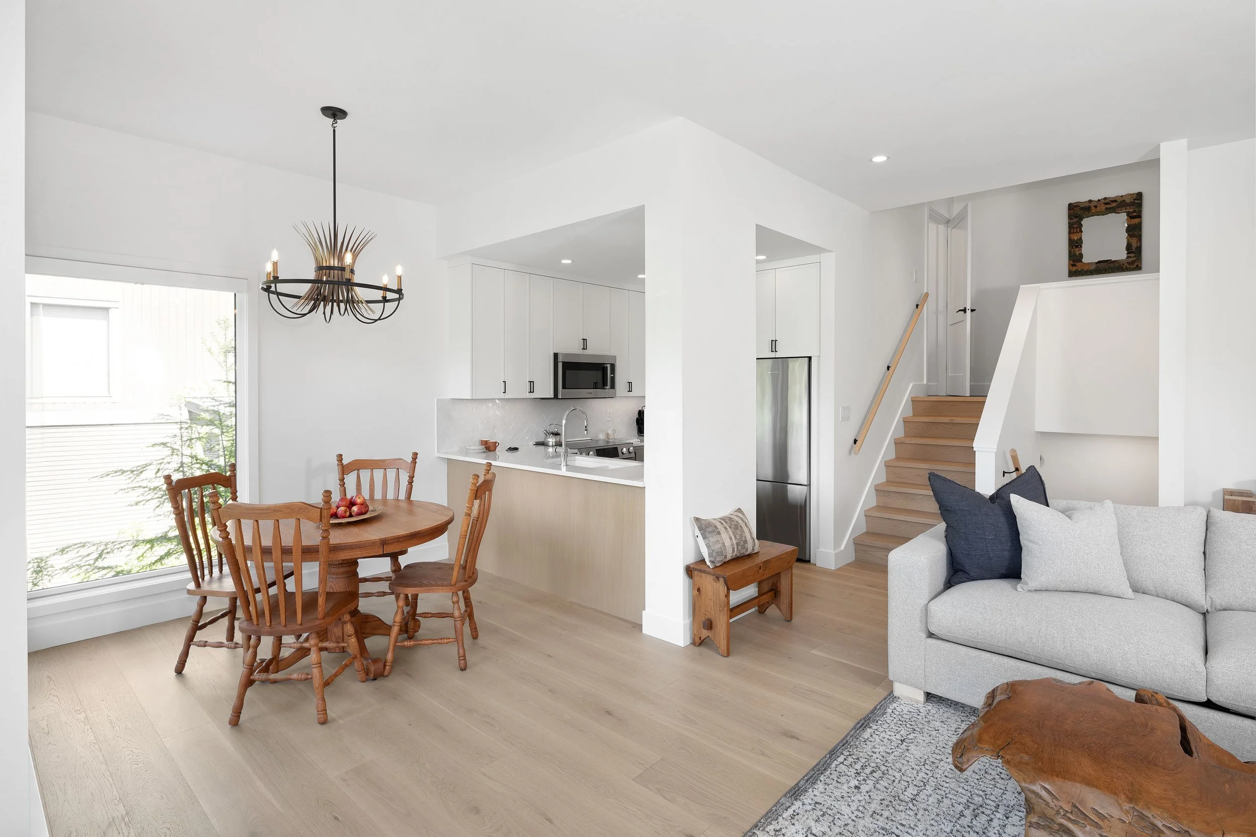 Open concept living and dining area with white walls, wooden flooring, a round dining table with four wooden chairs, a black and gold chandelier, a kitchen with white cabinets and stainless steel appliances, a staircase leading to upper floor, and a 