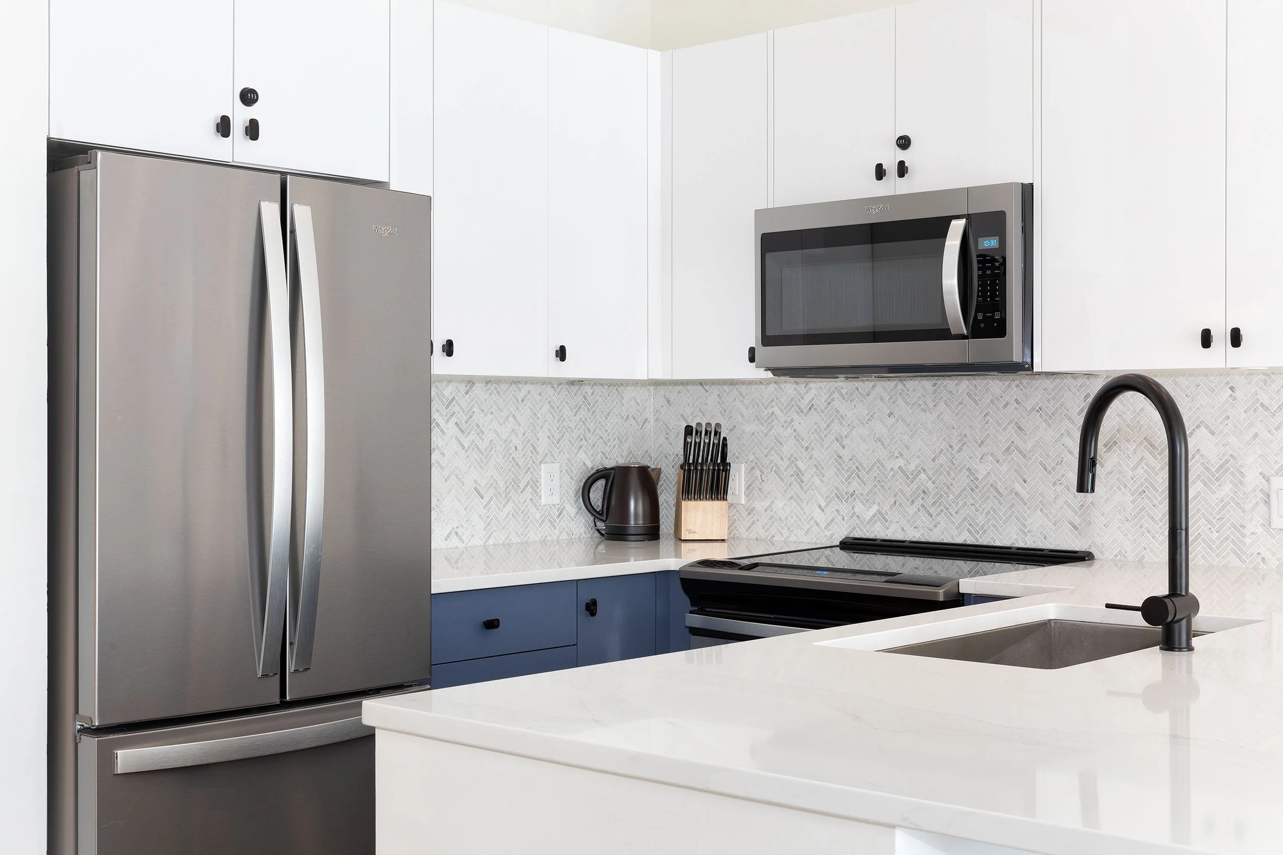 Modern kitchen with white cabinets, stainless steel refrigerator, microwave, countertop, black faucet, and backsplash.