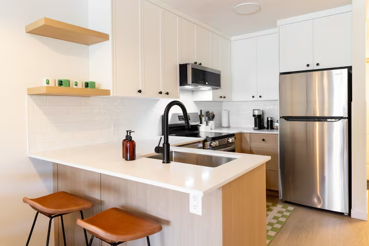 Modern kitchen with white cabinets, stainless steel refrigerator, black stove, black faucet, and wooden bar stools at the counter.