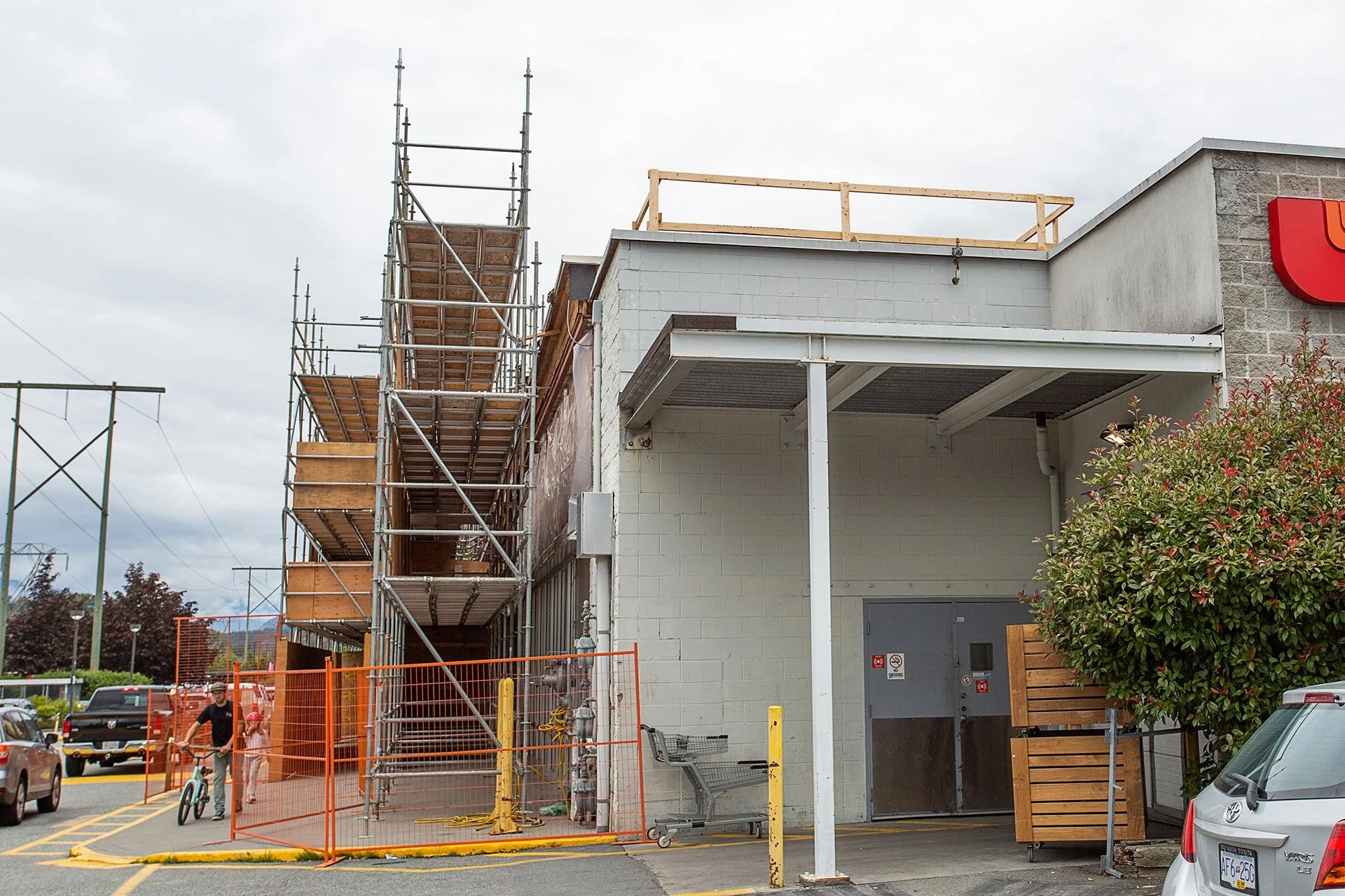 Construction site with scaffolding on the side of a building, workers and shopping carts in the area, and orange safety barriers surrounding the workspace.