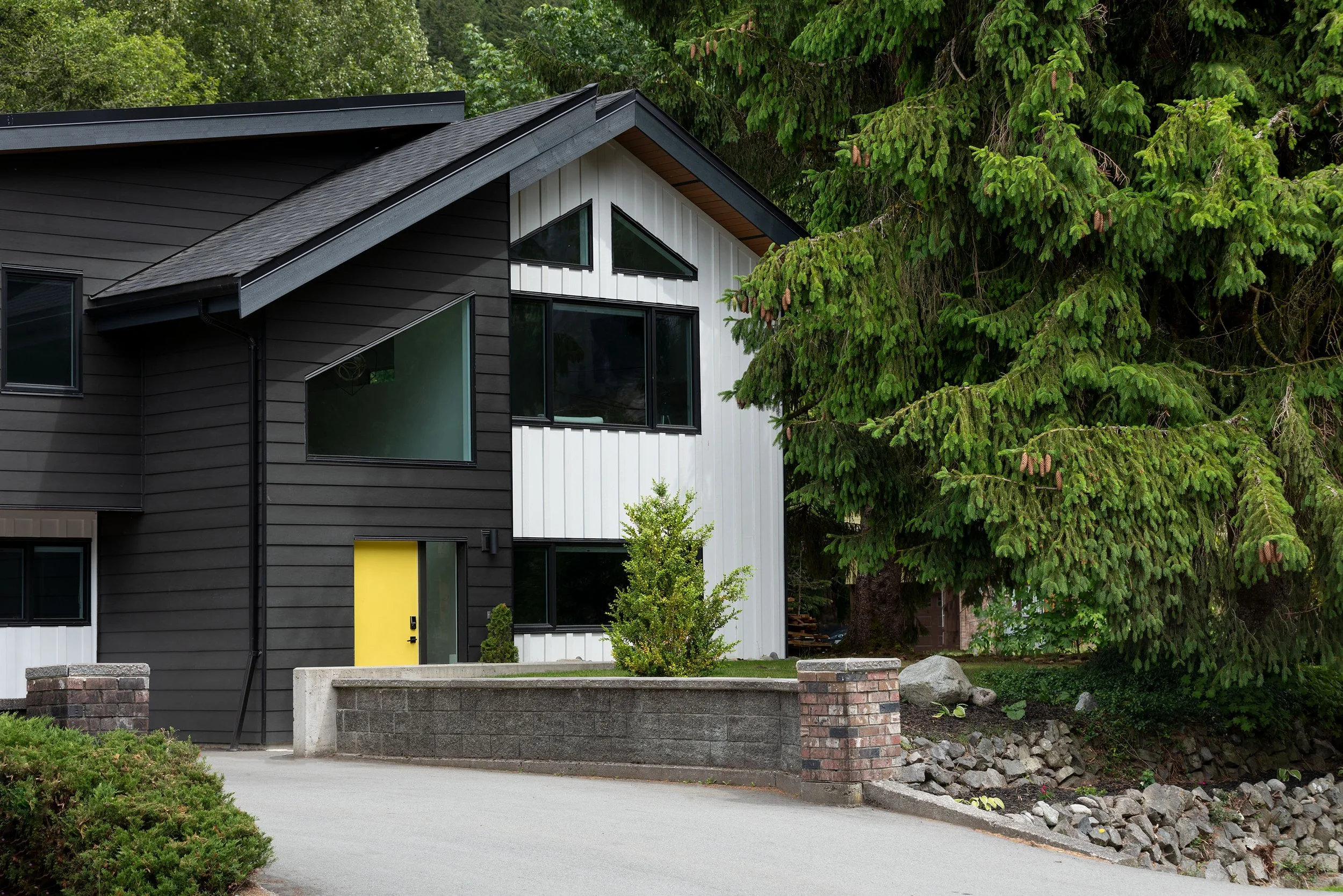 Modern two-story house with black and white exterior, yellow front door, large windows, surrounded by green trees and landscaped yard with rocks and shrubs.
