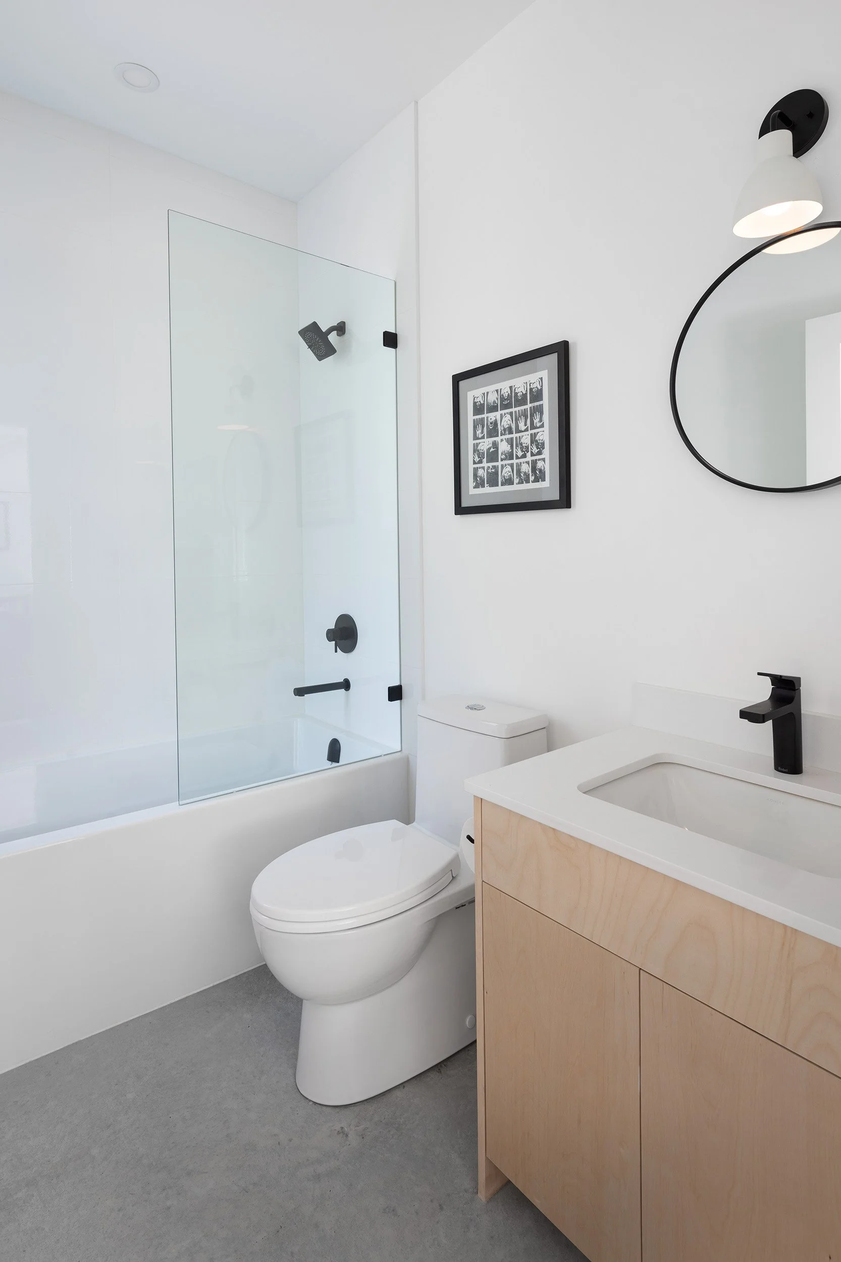 Modern minimalist bathroom with white walls, a glass shower enclosure, a white toilet, and a natural wood vanity with a sink. A black framed picture and a circular mirror with a black mount are on the wall, and a black wall-mounted light fixture is a